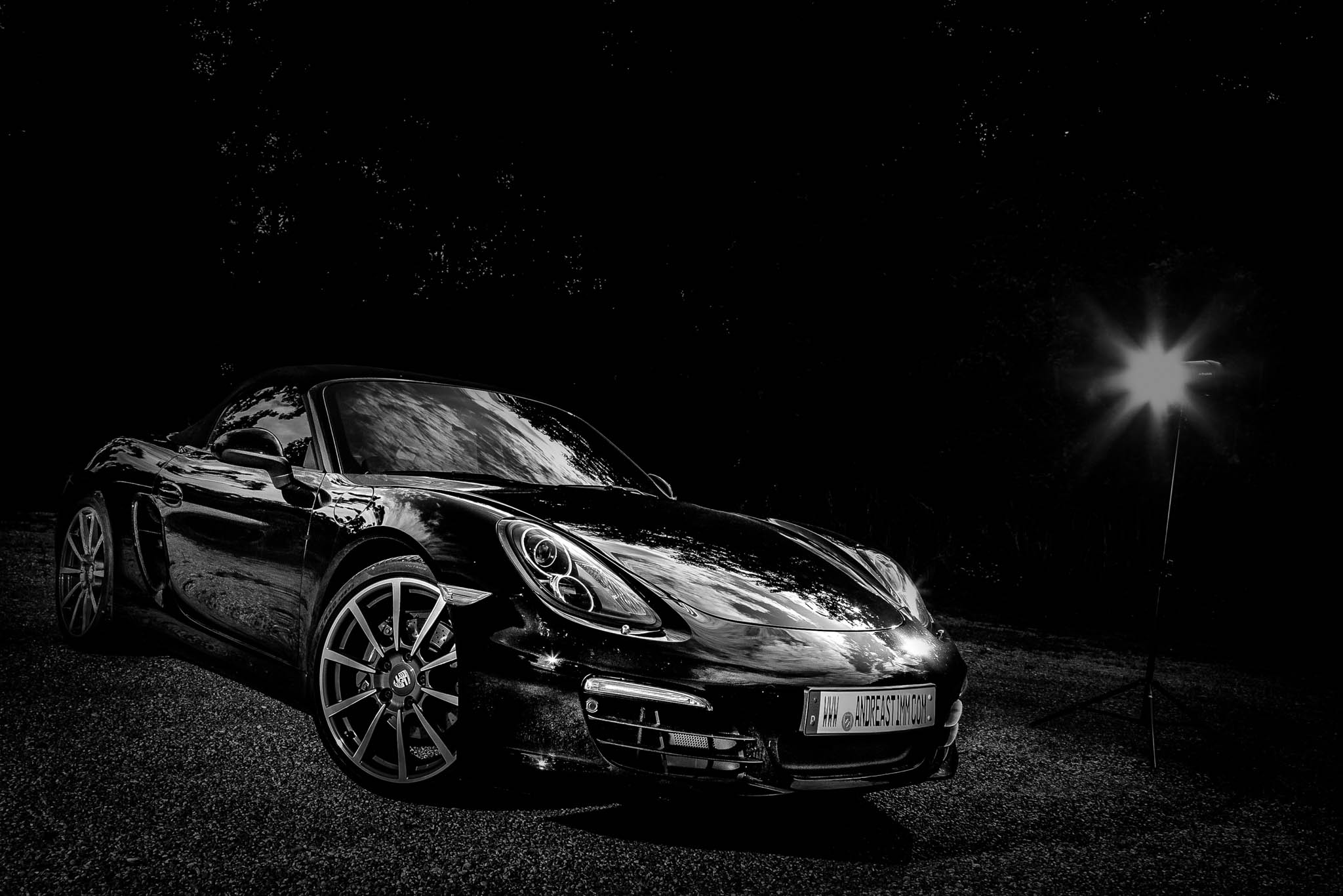 Black sports car illuminated by spotlight at night, reflecting trees on its shiny surface, parked on a gravel path.