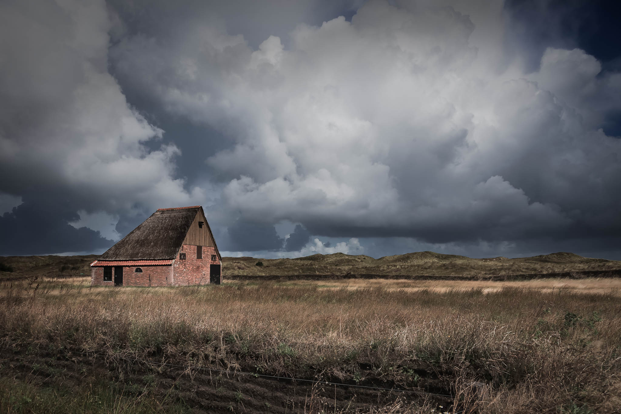 Rustic brick barn with a thatched roof amid vast fields under dramatic cloudy skies.