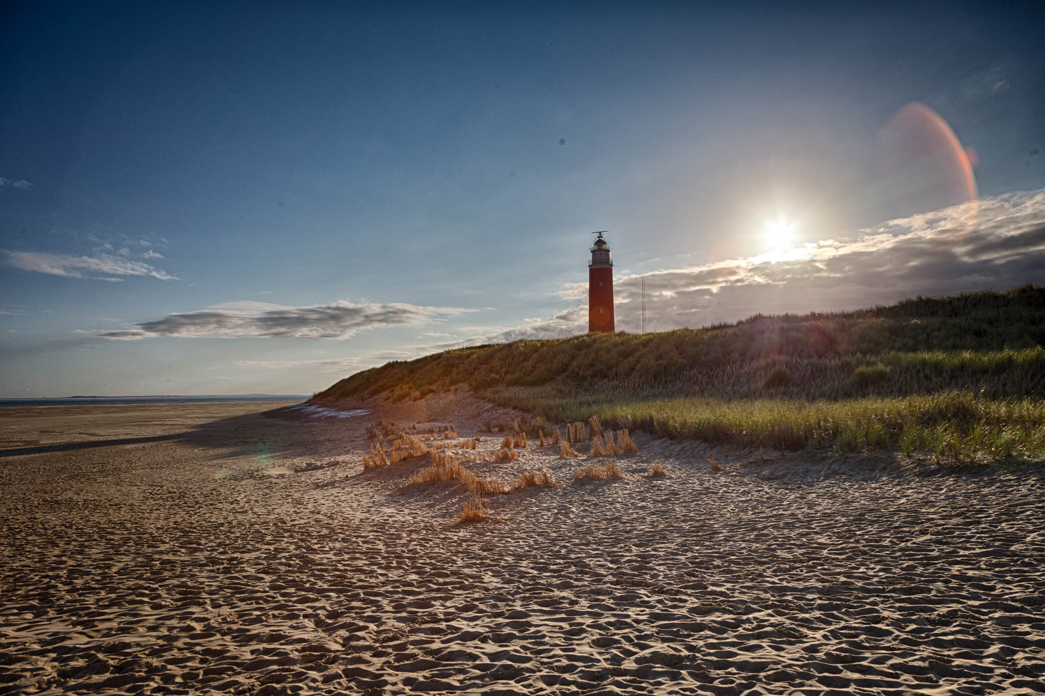 Sunny beach landscape with a red lighthouse on a grassy dune under a clear blue sky.