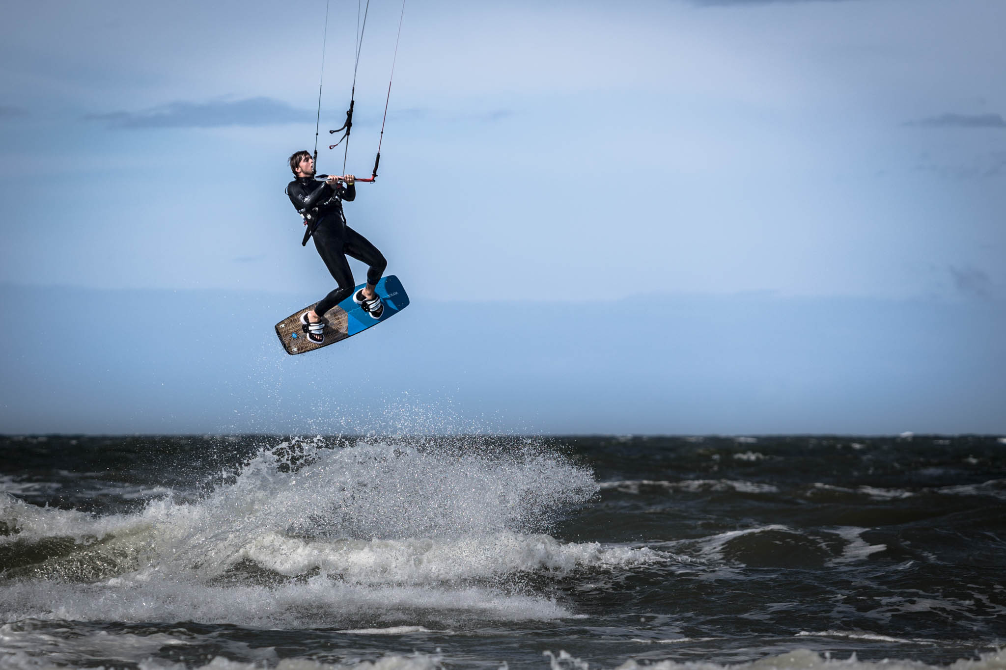 Kitesurfer in mid-air above the ocean, performing a jump against the blue sky with waves crashing below.