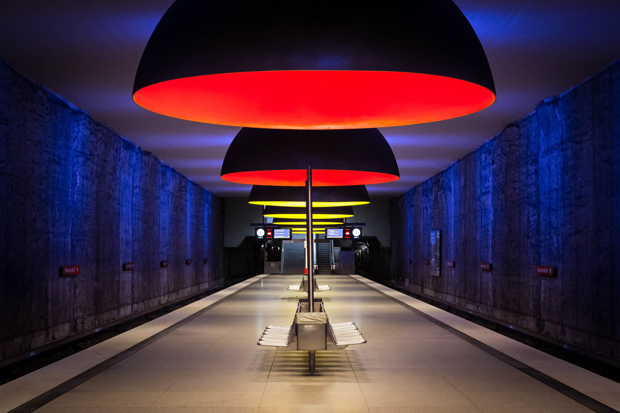 Modern subway station with vibrant red and yellow ceiling lights and blue walls, emphasizing symmetry and design.