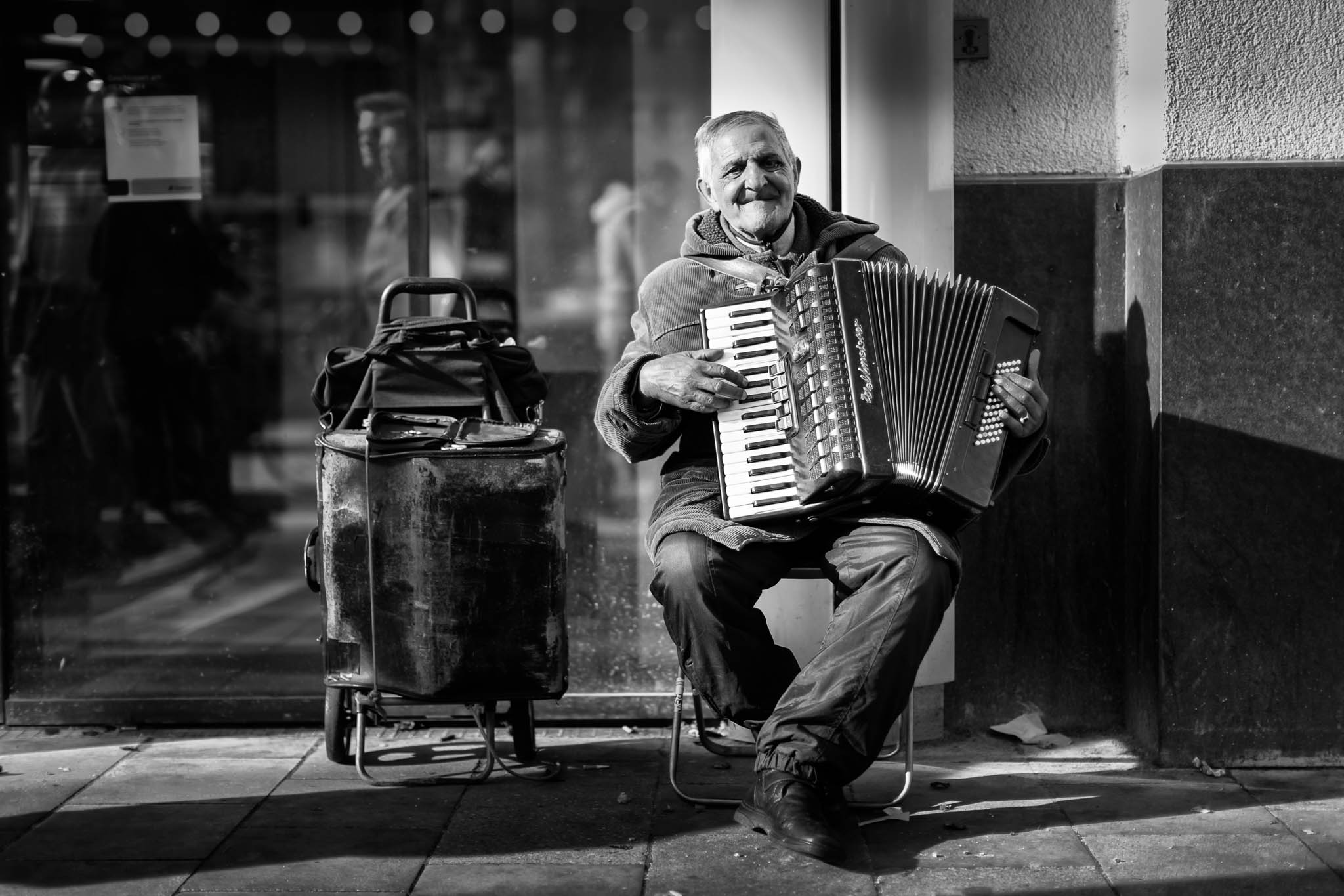 Elderly man playing accordion on urban street corner with suitcase beside him, conveying a street musician's serene moment.