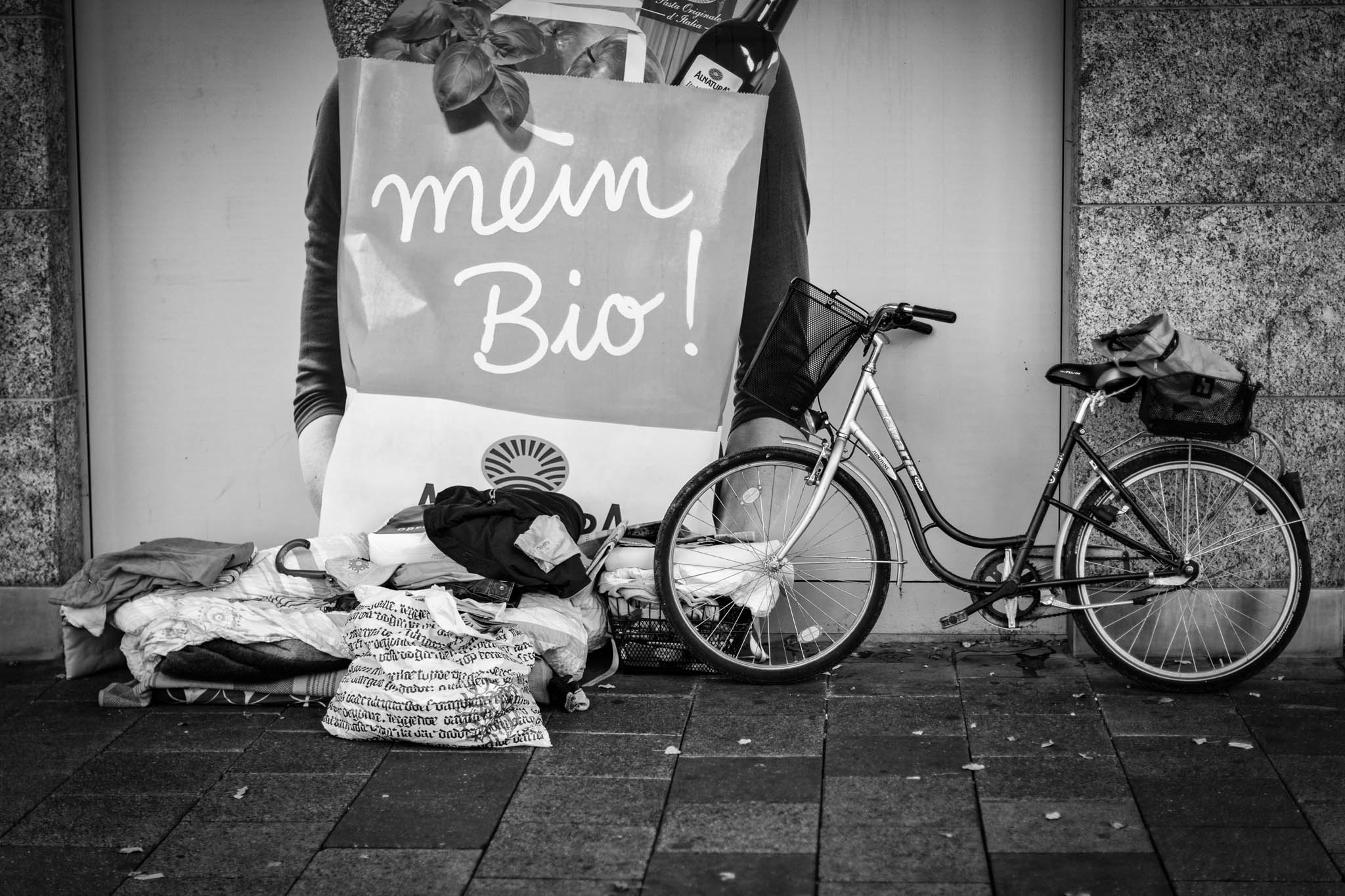 Bicycle parked by wall with grocery bag billboard, surrounded by blankets and clothing on the ground in a black and white scene.