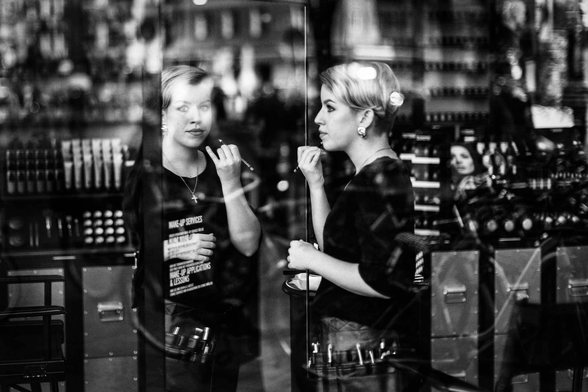 Woman applying makeup in a cosmetics store, reflected in the glass display, surrounded by beauty products. Black and white.