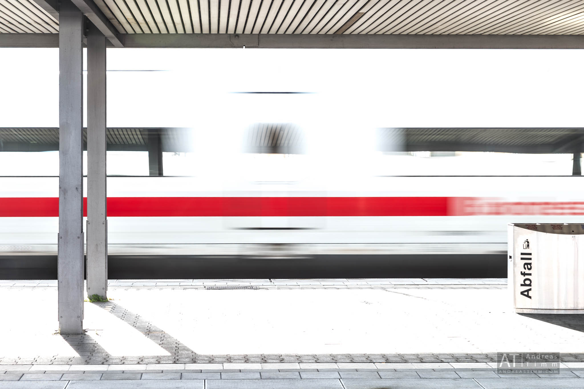 Blurred high-speed train passing through a modern station platform with a trash bin labeled Abfall.