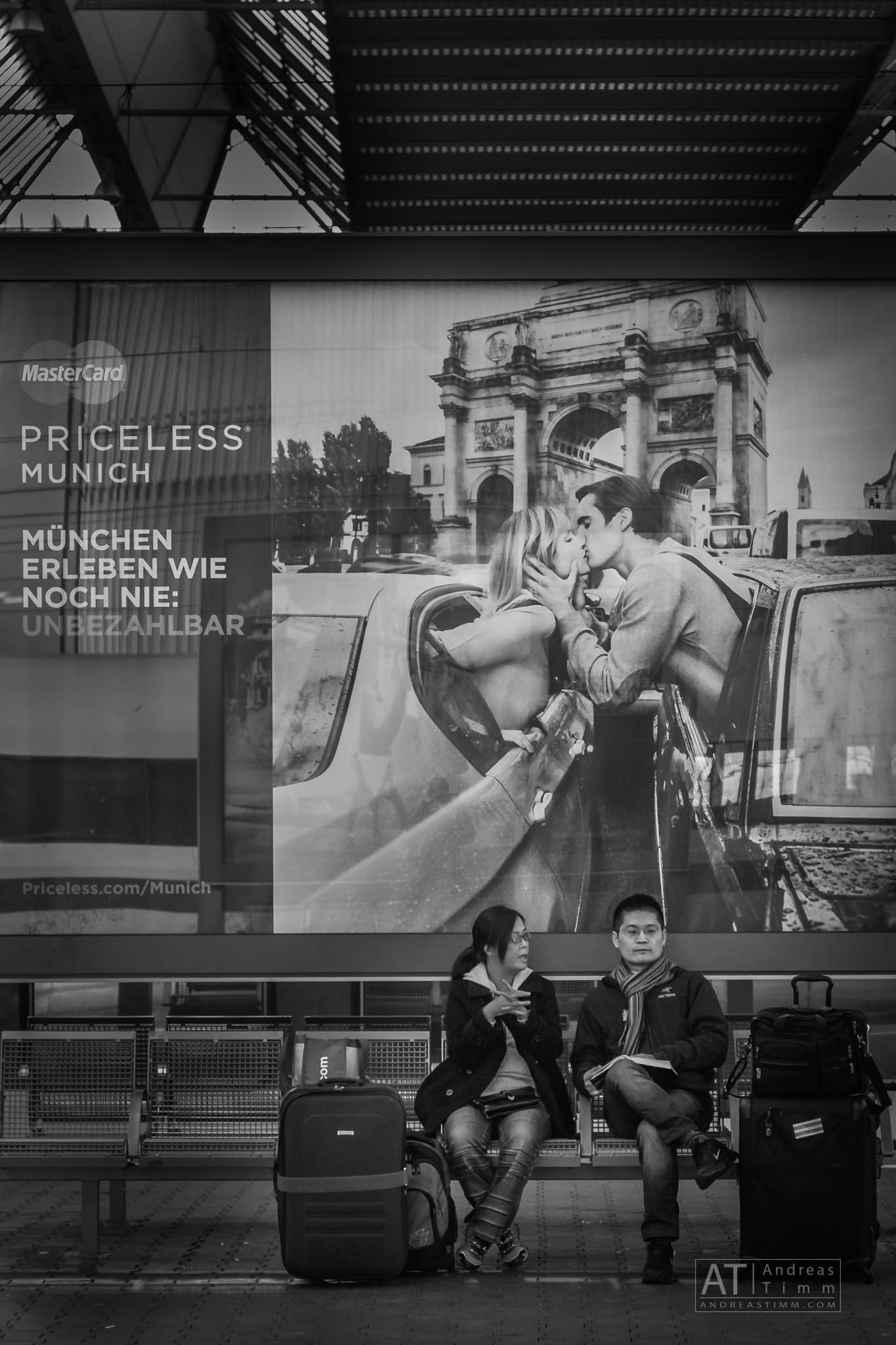 Travelers sit under a large advert of a couple kissing near Munich's iconic landmark at a station.