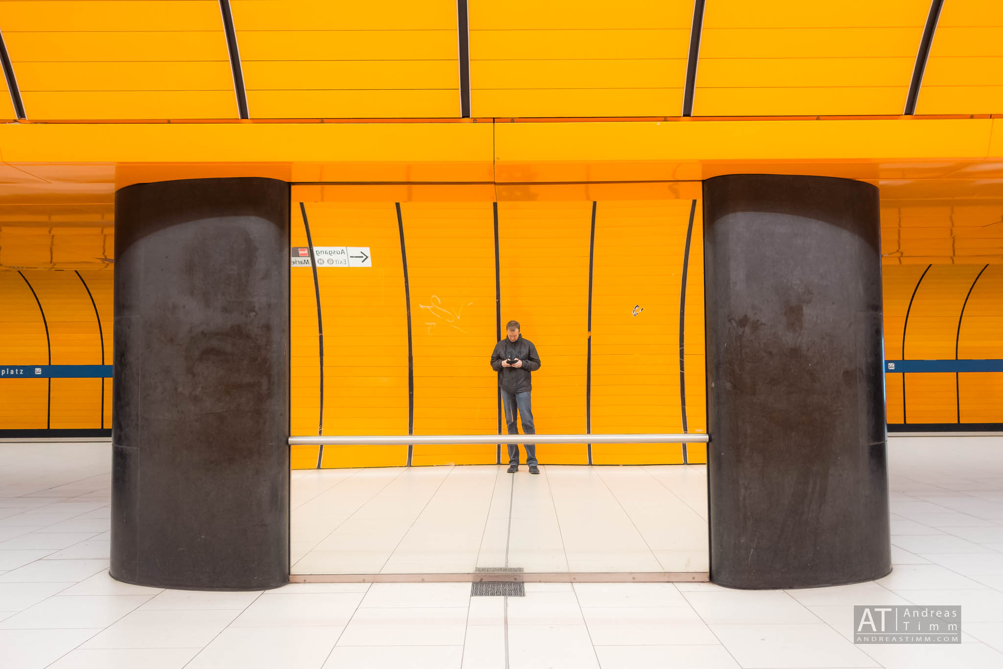 Man standing in bright orange subway station, looking at phone, next to large black pillars.