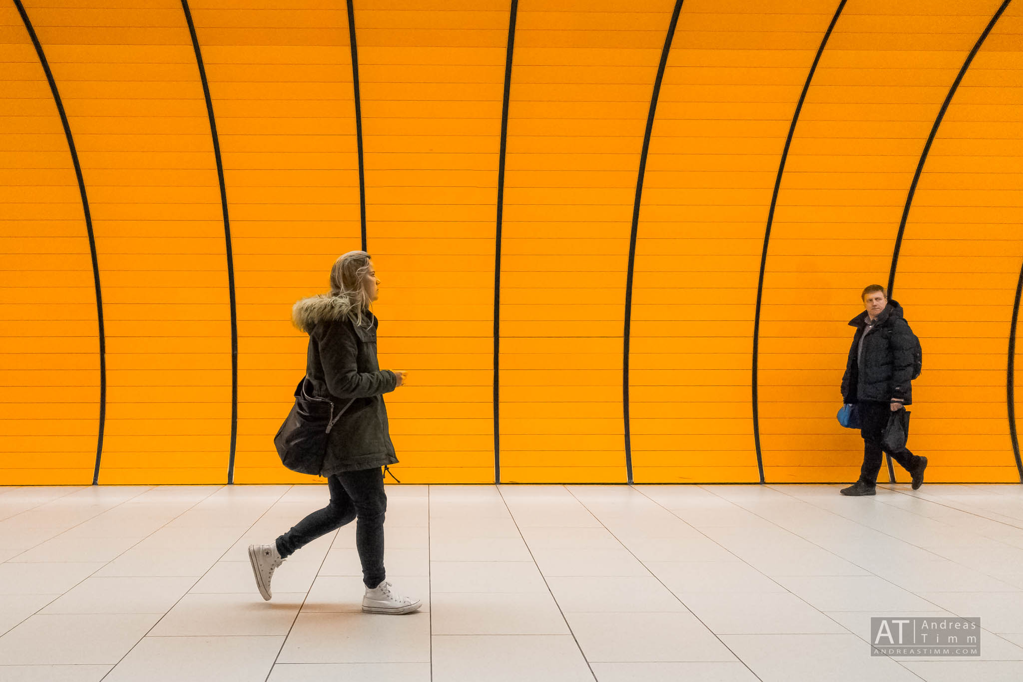 Two people walking in a subway station with bright orange curved walls and white tiled floor.