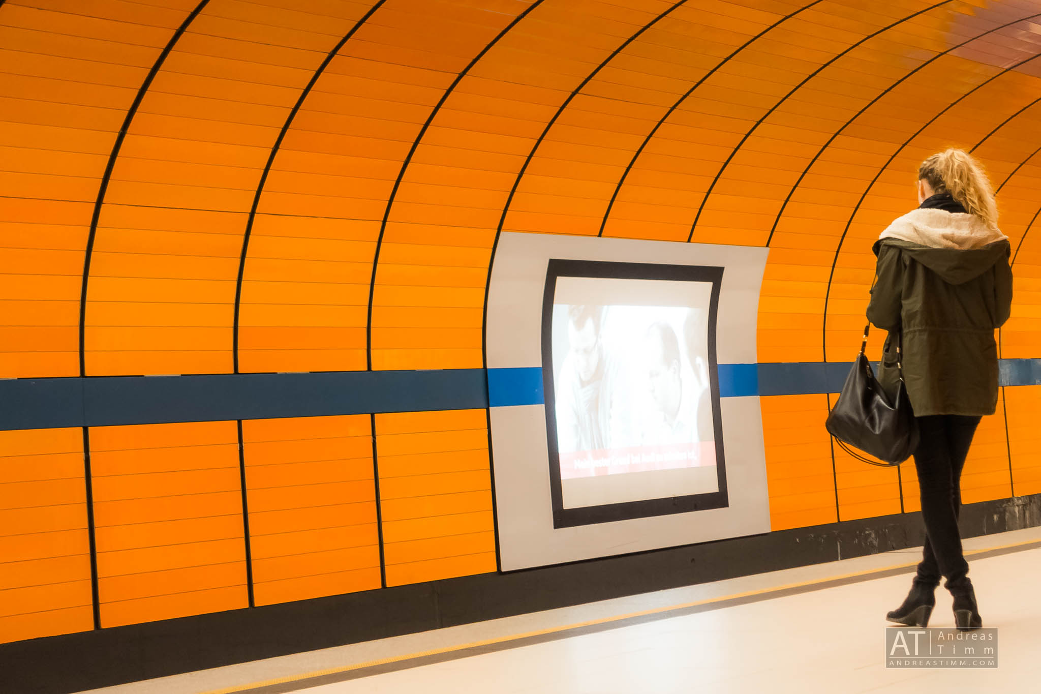 Person walking in an orange subway tunnel with a curved wall and digital screen display.