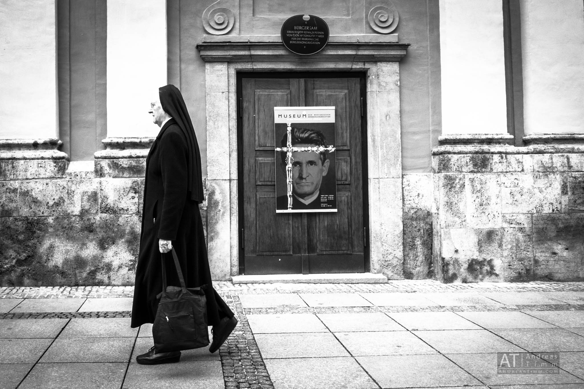 Nun walking past a museum door with a poster, in a black and white streetscape scene.