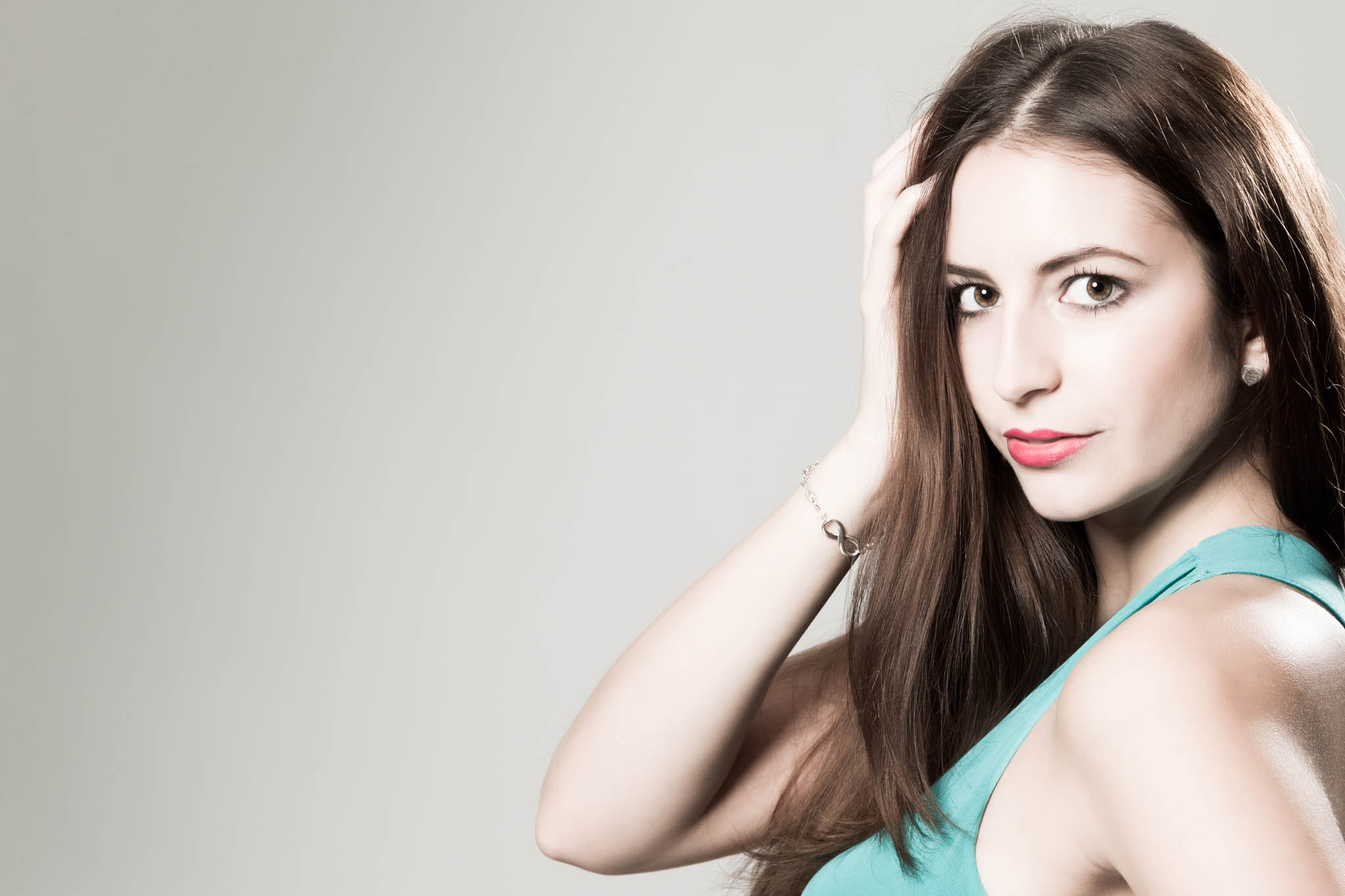 Woman with long brown hair in teal top, touching her head, against a light gray background.