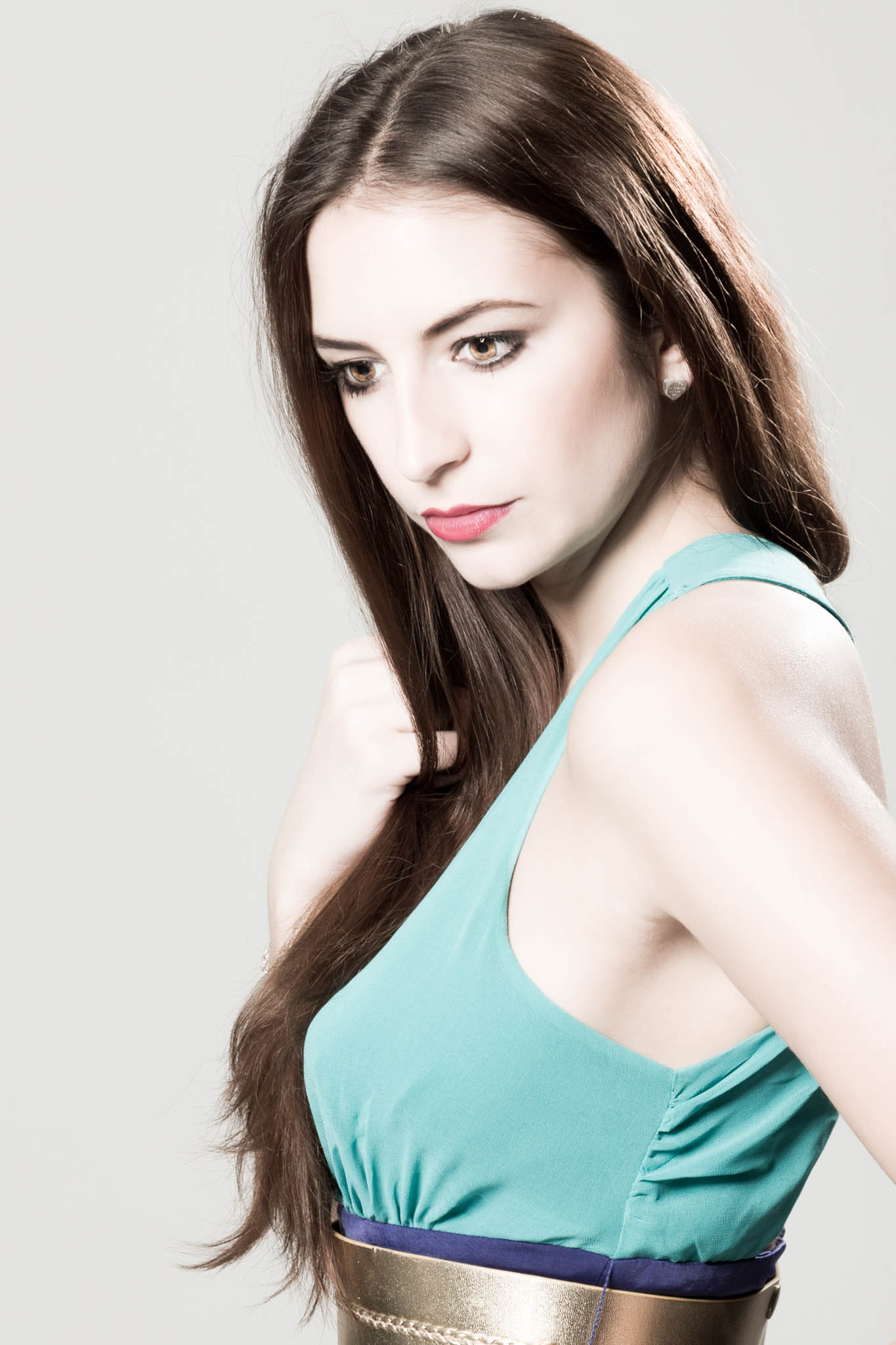 Woman with long brown hair wearing a teal dress and gold belt, looking down thoughtfully against a light gray background.