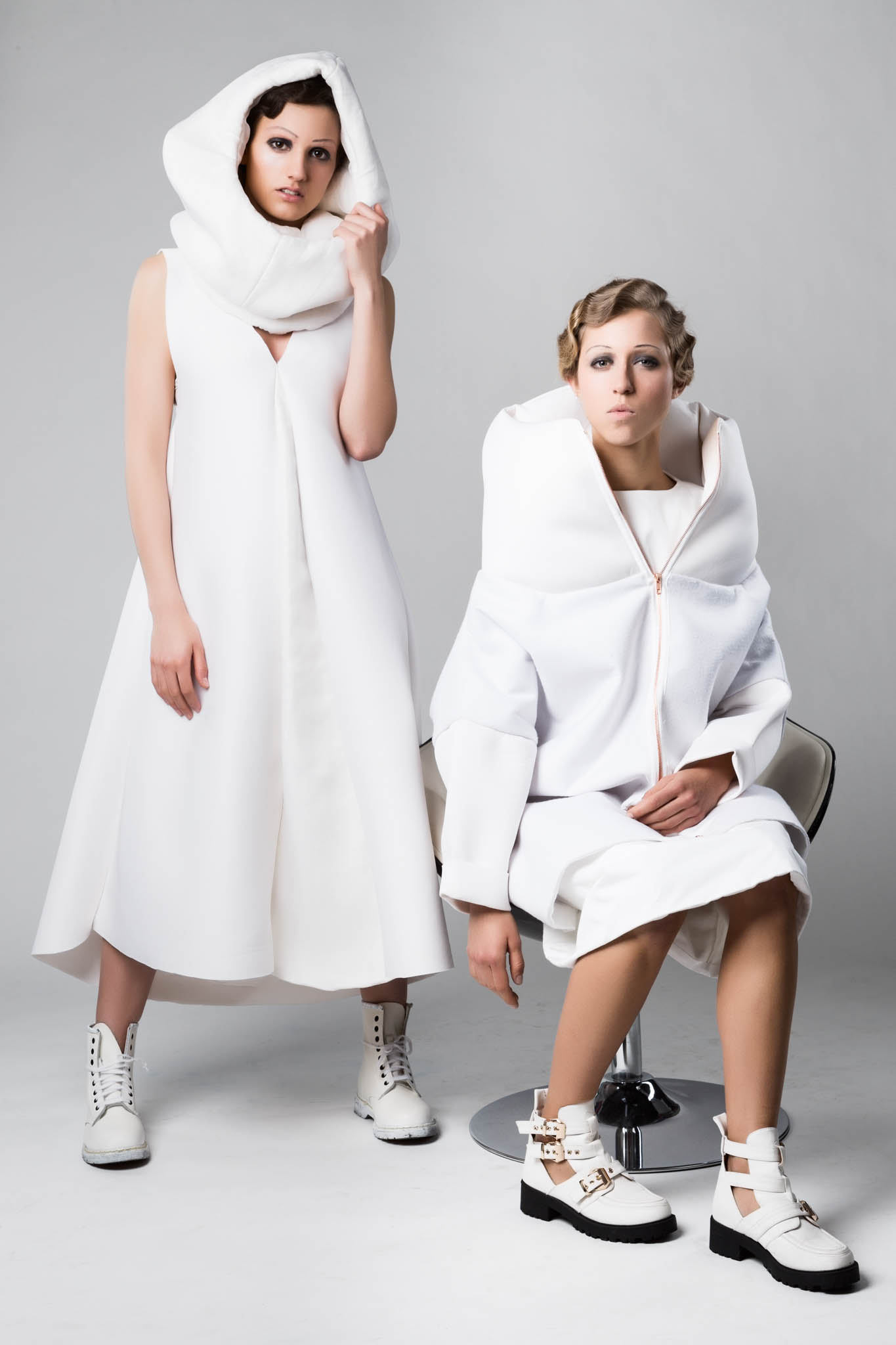 Two models in creative, white avant-garde fashion outfits with unique hoods and boots against a neutral background.