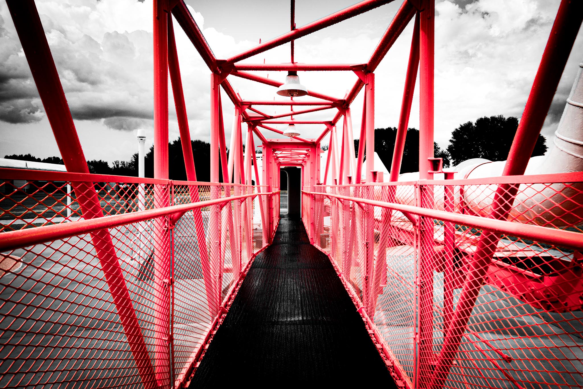 Red metal bridge with crisscross pattern against cloudy sky, leading to a narrow passage.
