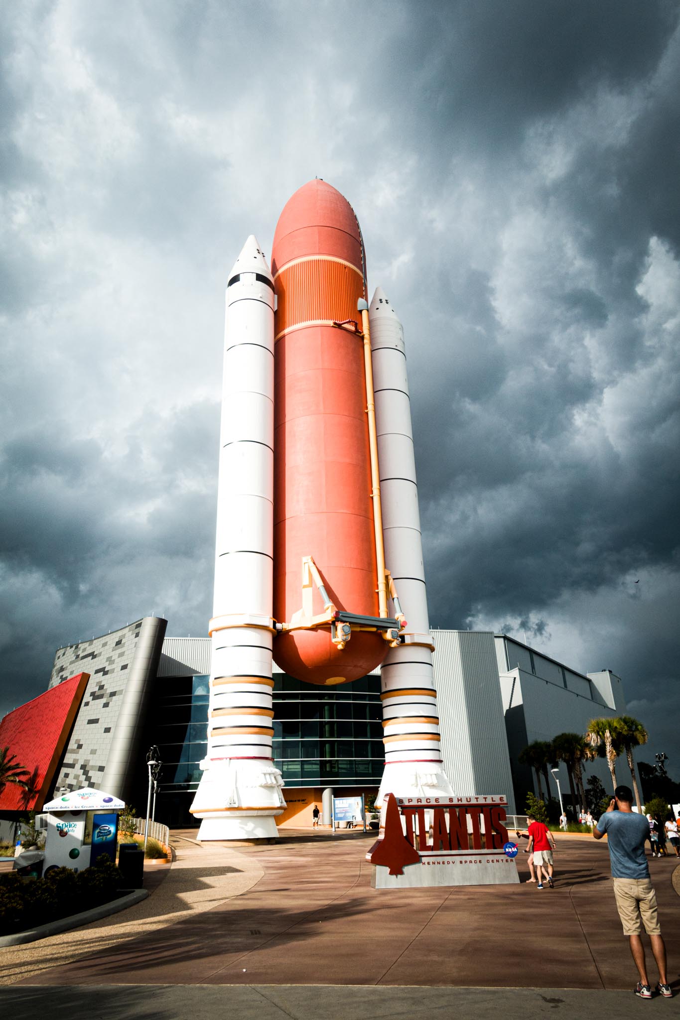 Space shuttle replica at Kennedy Space Center with dramatic cloudy sky, visitors at entrance.