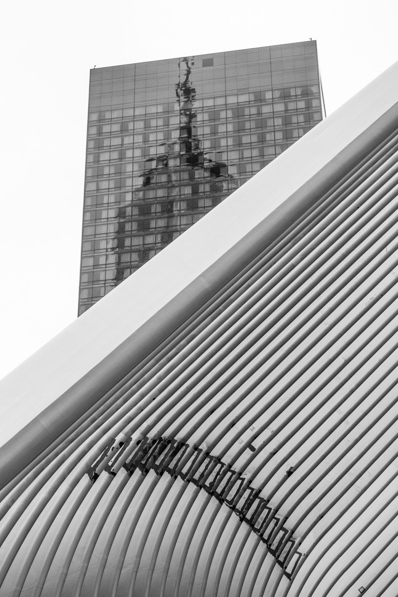 Curved architectural structure with a building reflected in glass facade above; black and white urban scene.