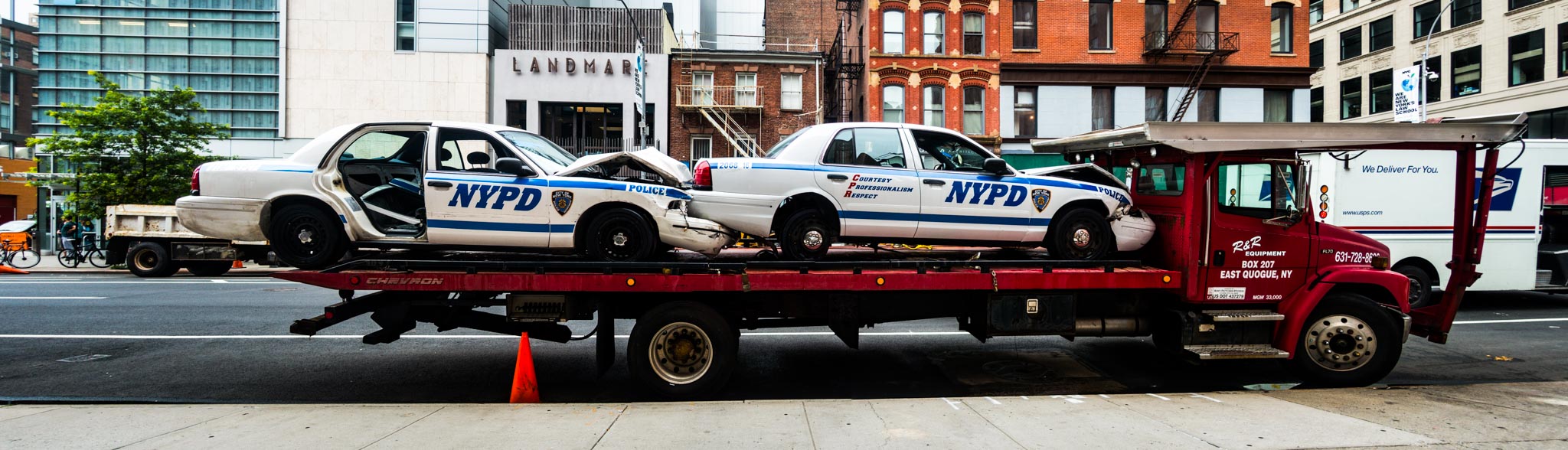 Tow truck carrying two damaged NYPD police cars on a city street.