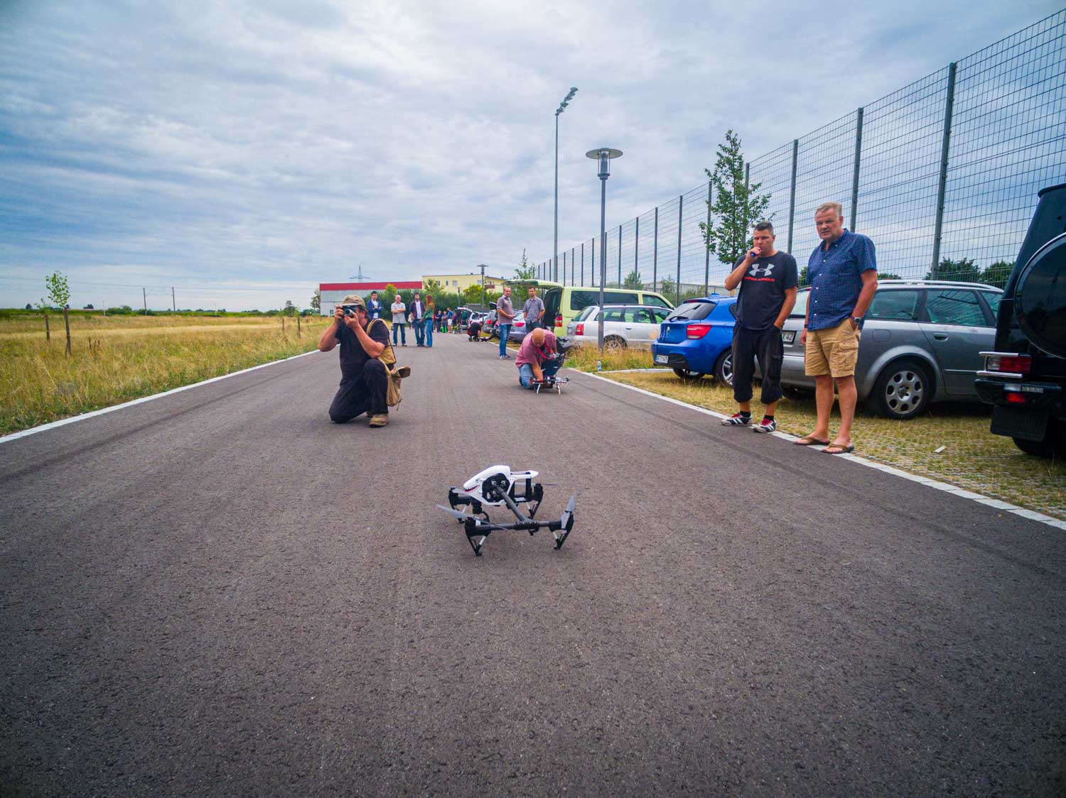 Man photographing a small drone on an outdoor road with people and parked cars in the background.