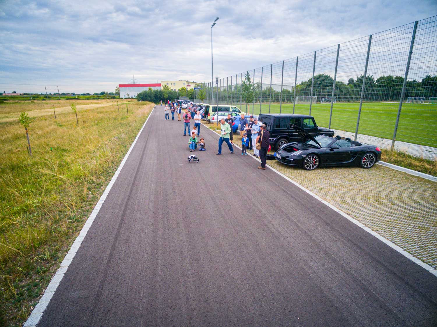 People gathered near parked cars on a rural road beside fields and a soccer field, with a toy car on the road.