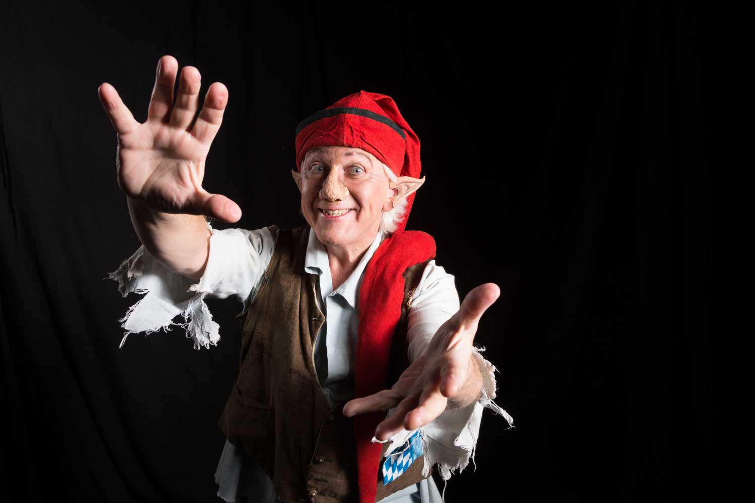 Smiling man dressed as an elf with red hat and outstretched arms against a black background.