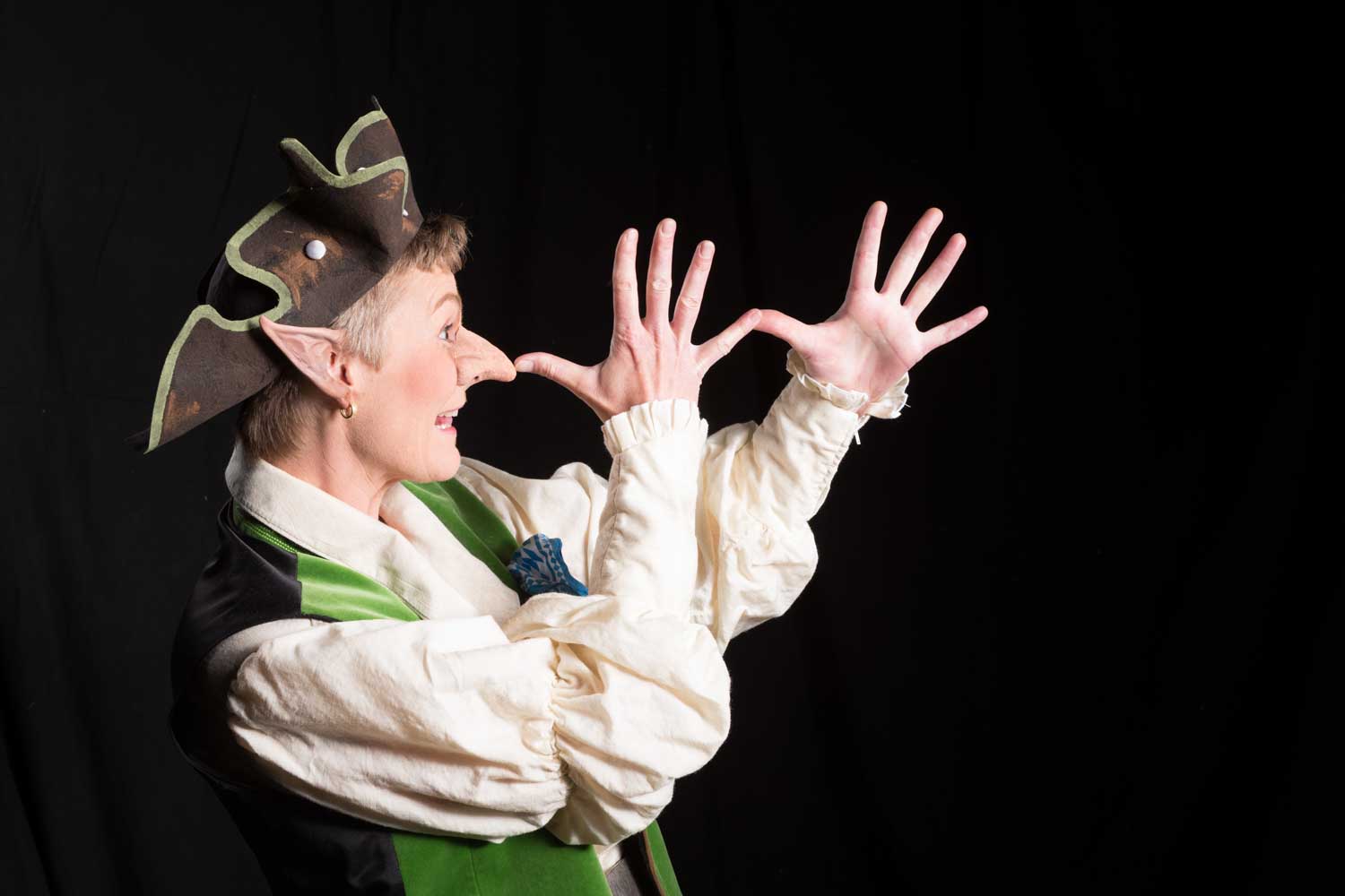 Person in whimsical costume with pointy hat, long nose, and expressive hands against a black background.