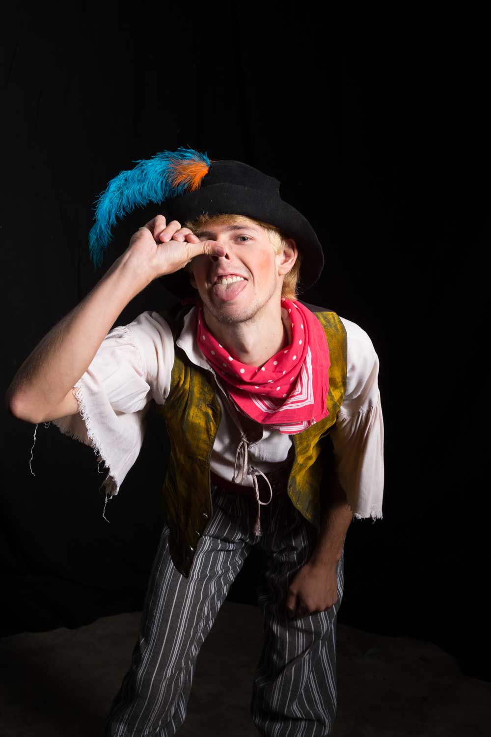 Costumed man with feathered hat and striped pants making a playful face, sticking out tongue with thumb on nose.