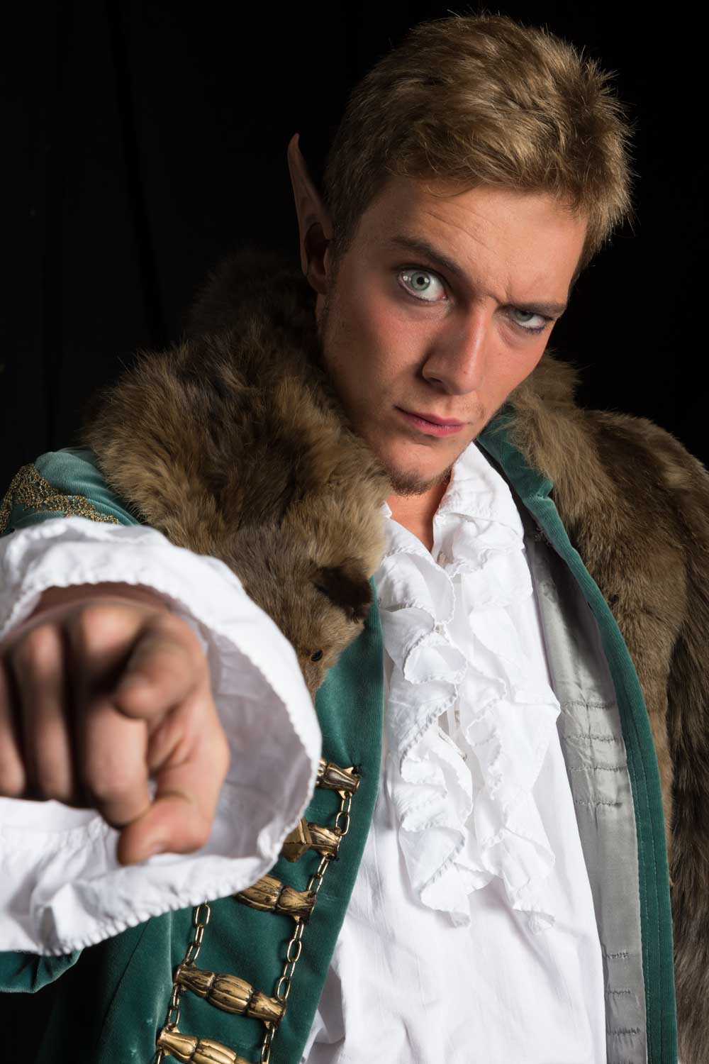 Fantasy character in fur cloak pointing forward, wearing white ruffled shirt, intense gaze, and elvish ears.