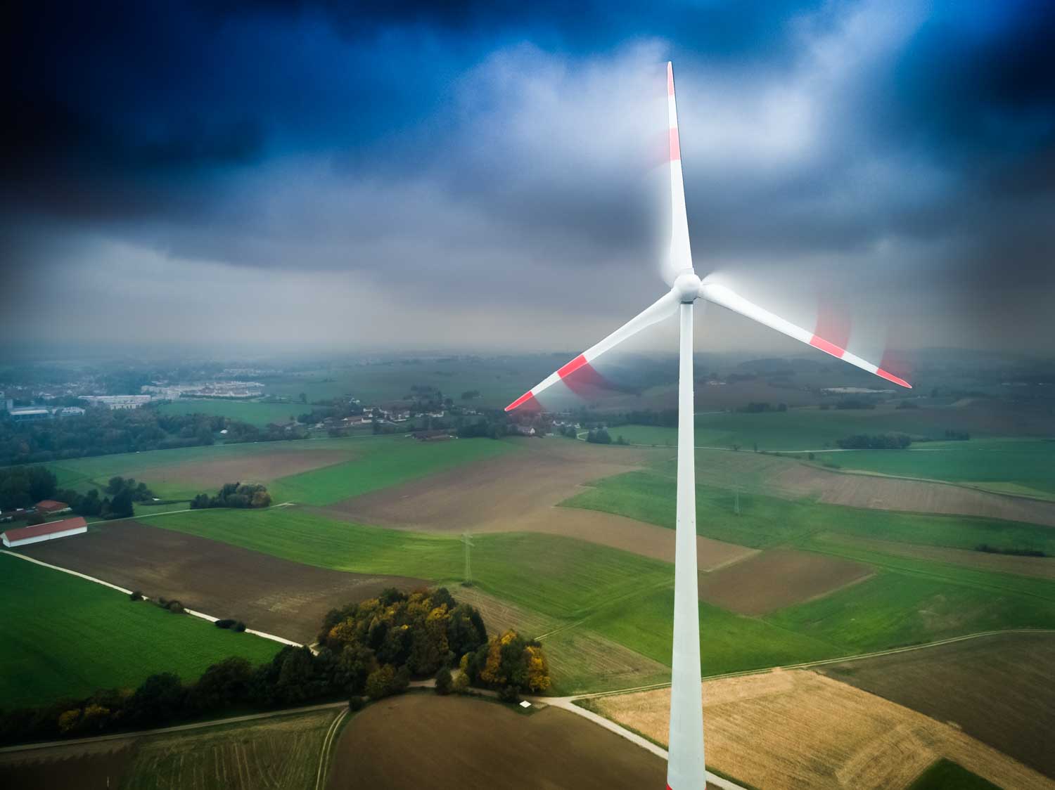 Wind turbine spinning over vast green and brown farmland under a cloudy sky.