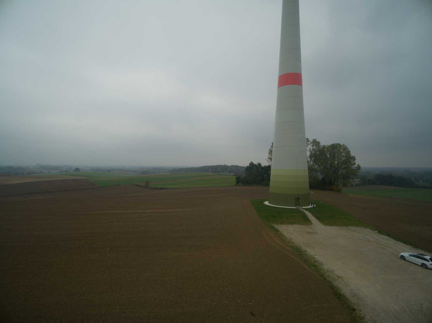 Wind turbine tower on a cloudy day, surrounded by fields, with a parked car nearby.