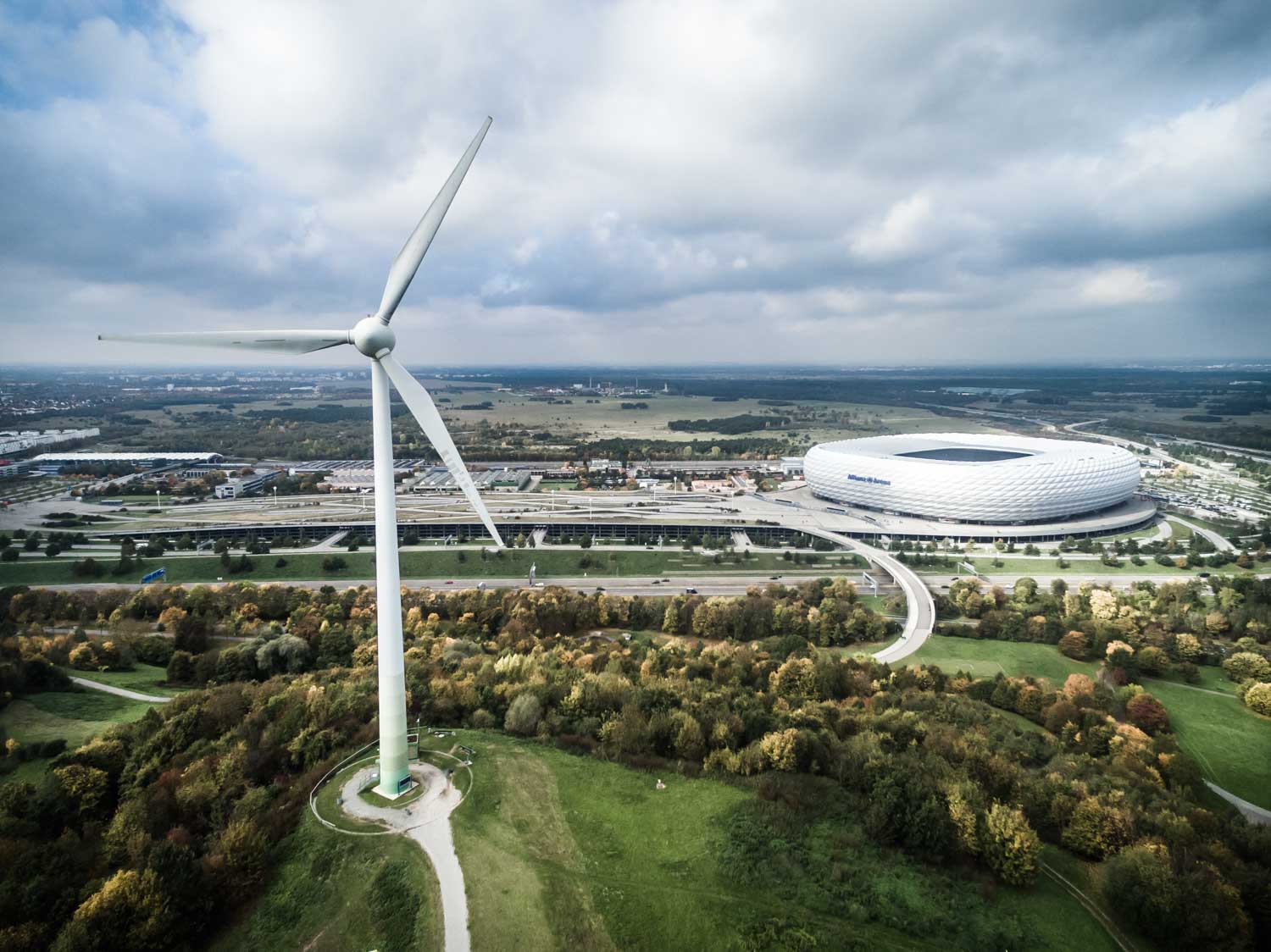 Aerial view of a wind turbine near a large stadium surrounded by green fields and roads on a cloudy day.