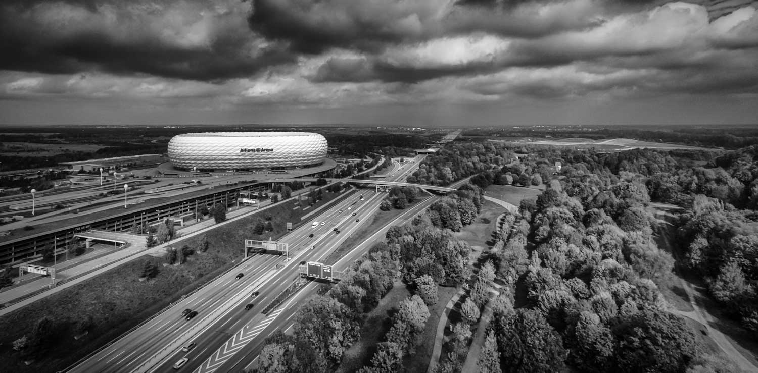 Aerial view of stadium near highway surrounded by trees under dramatic cloudy sky in black and white.