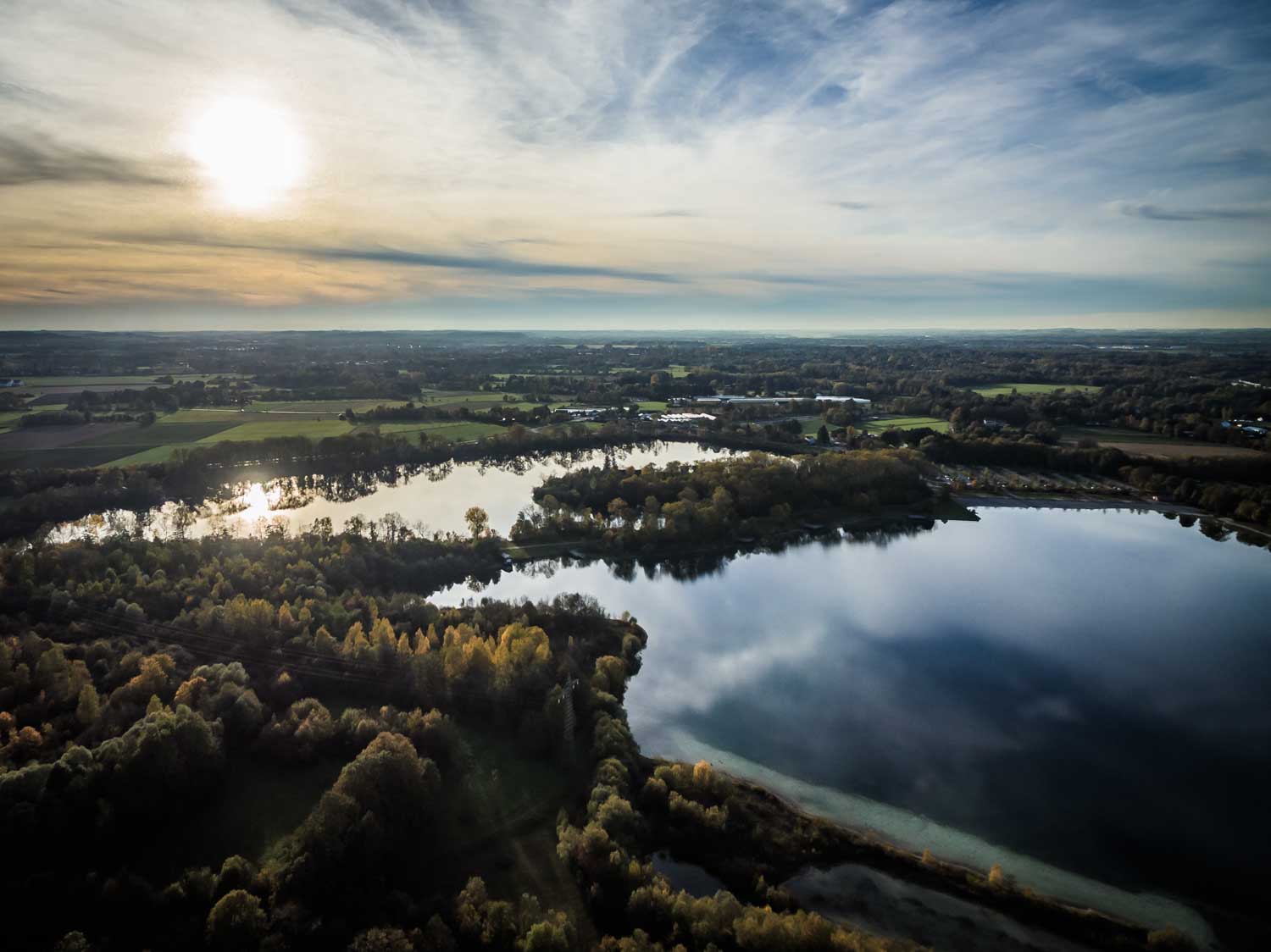Aerial view of a serene lake reflecting the cloudy sky, surrounded by forests and fields under a setting sun.