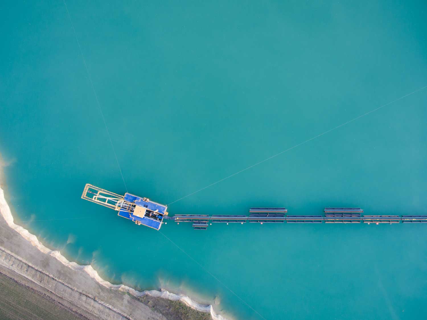 Aerial view of a blue dredging barge on teal water, with pipes extending to the shoreline.