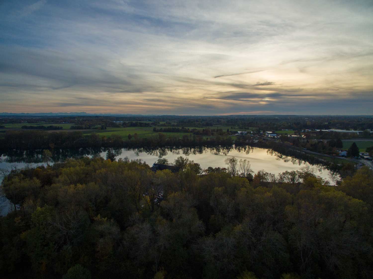 Aerial view of a serene lake surrounded by lush trees and open fields under a cloudy, dusk sky.