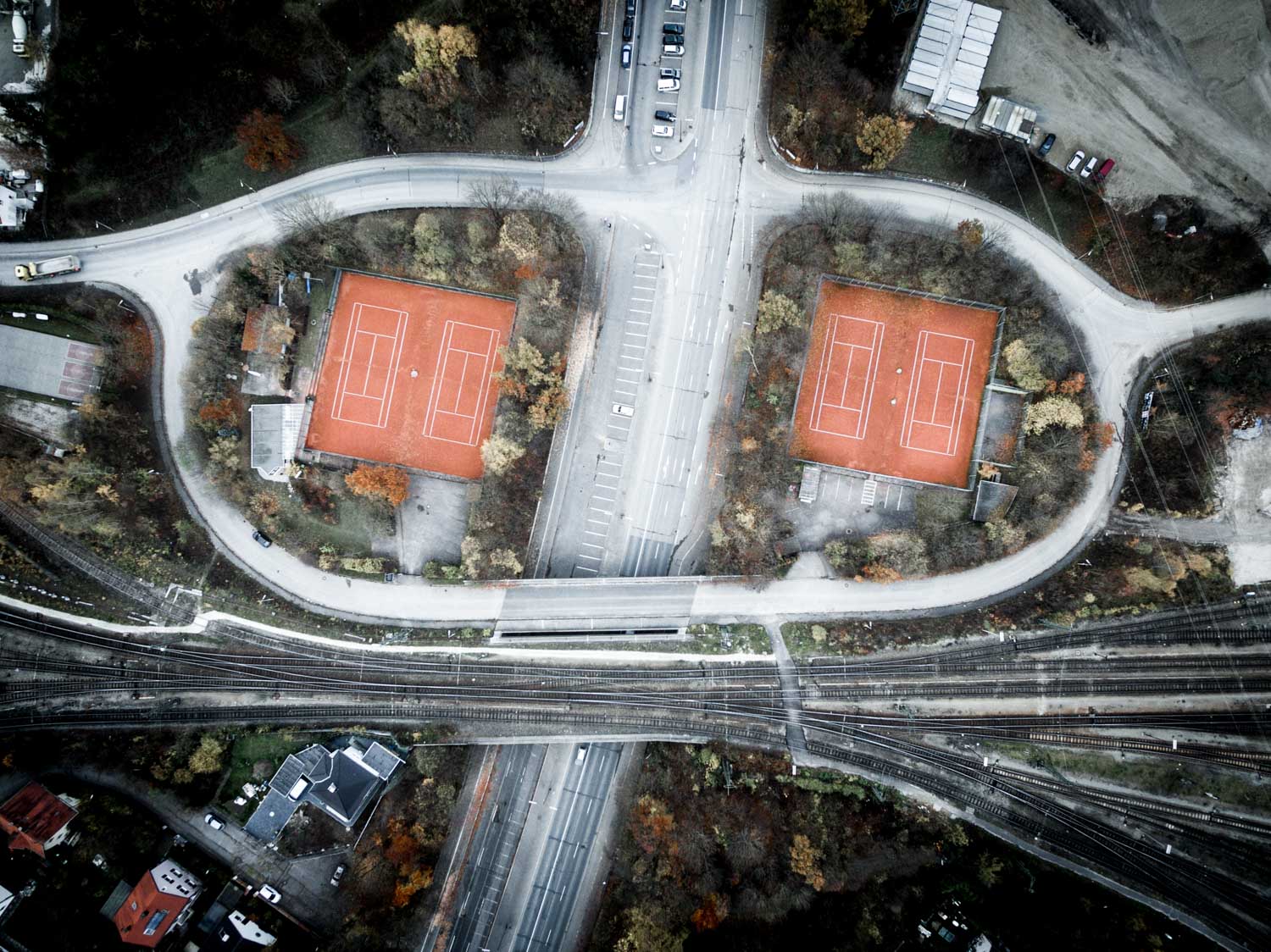 Aerial view of two adjacent red clay tennis courts surrounded by trees and roads, near a railway line.