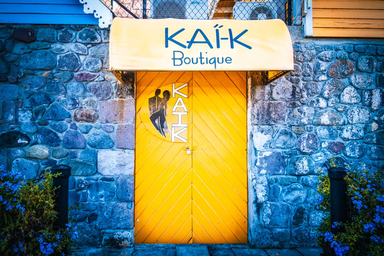 Yellow door with Kaï-K Boutique awning against stone wall, featuring a silhouette logo and surrounded by flowers.