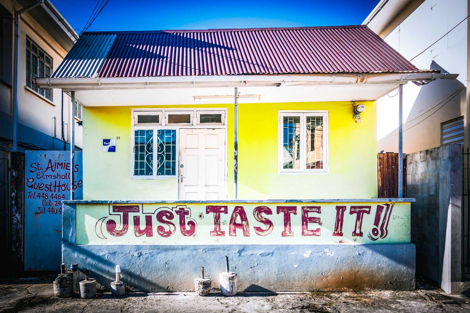 Yellow building with white door, red text Just TASTE IT! on wall, beside guesthouse sign. Bright sunny day.