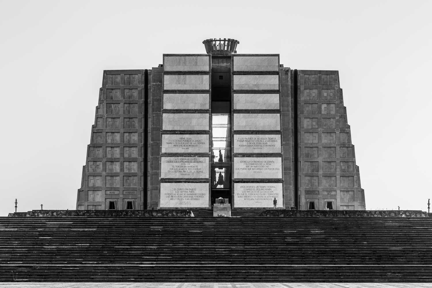 Black and white image of stone monument with large steps and engraved text, surrounded by an open, empty plaza.