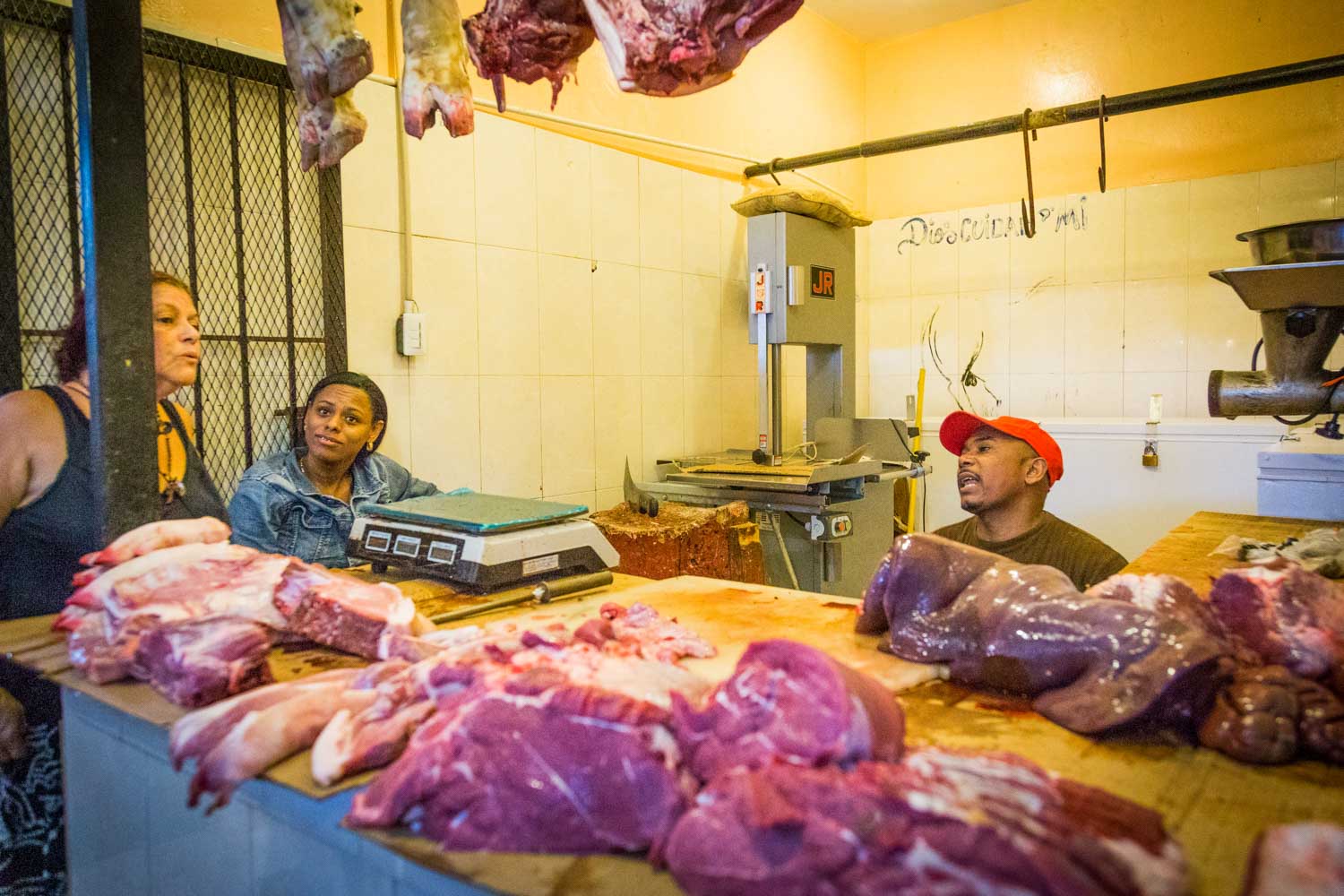 Butcher shop with fresh meat cuts on display, two women and a man engaged in conversation behind the counter.