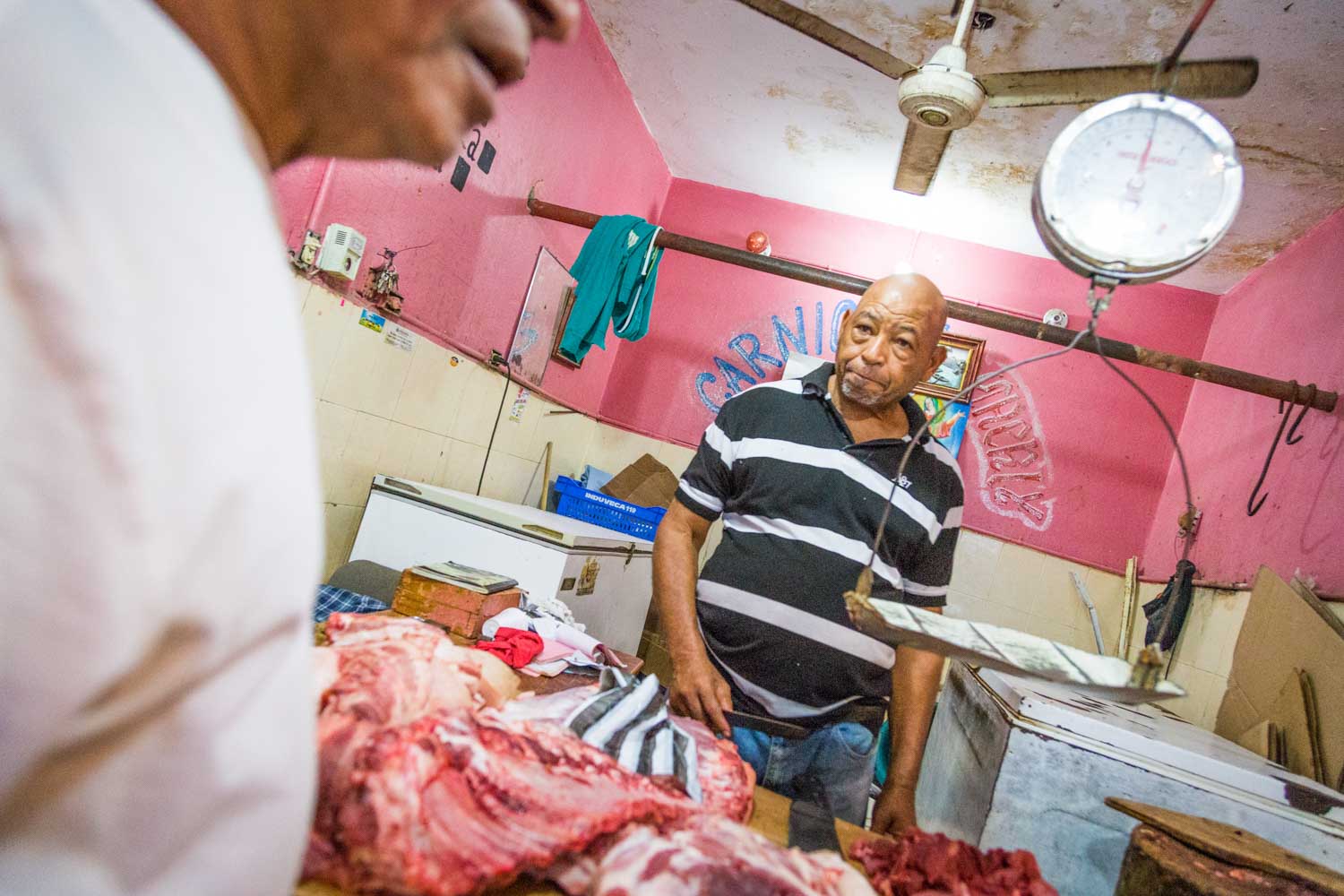 Butcher in striped shirt standing beside meat counter in a pink-walled shop with a ceiling fan and hanging scale.