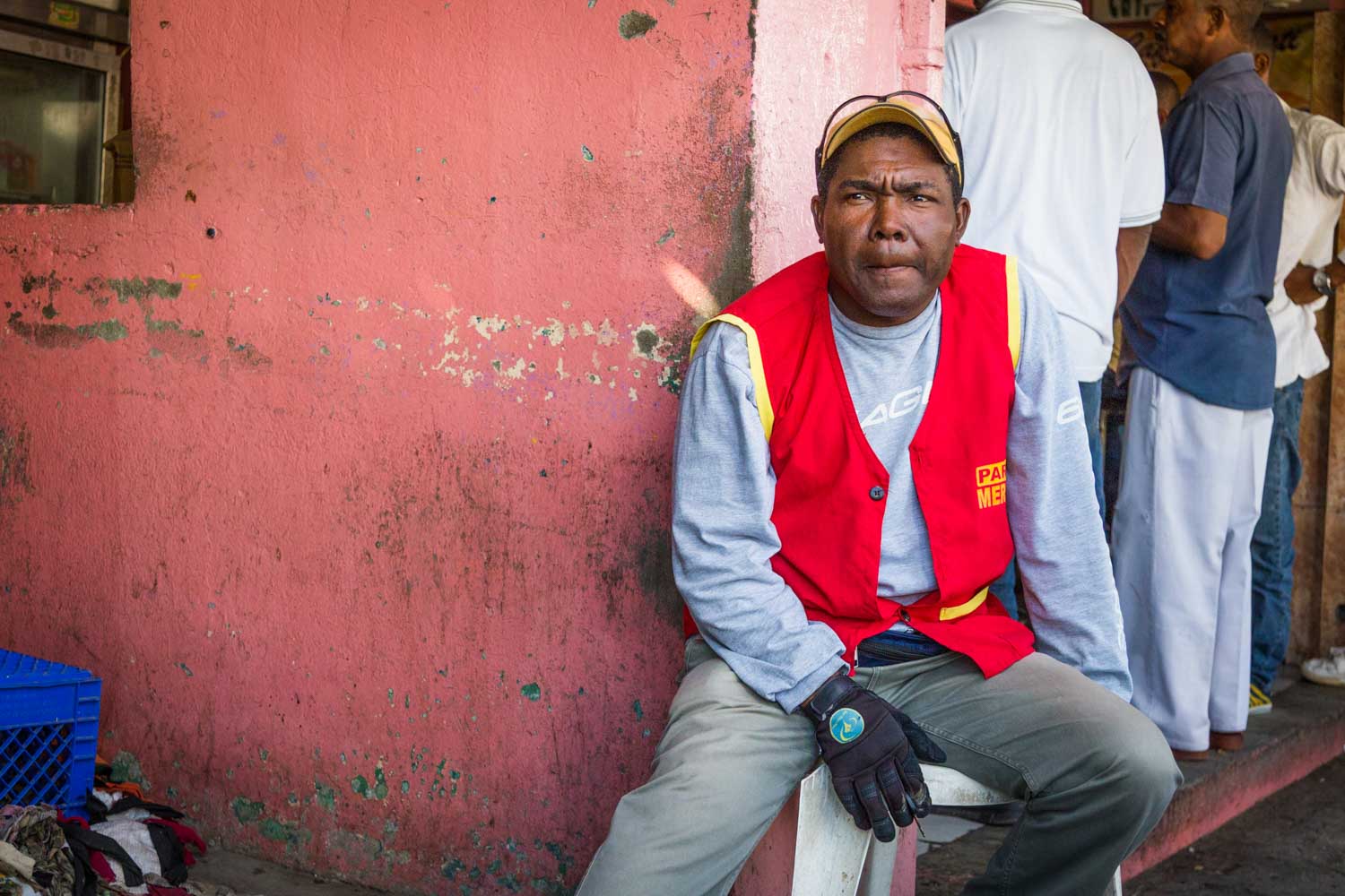 Man in a red vest sitting by a pink wall, wearing a cap and gloves, with people standing behind him.