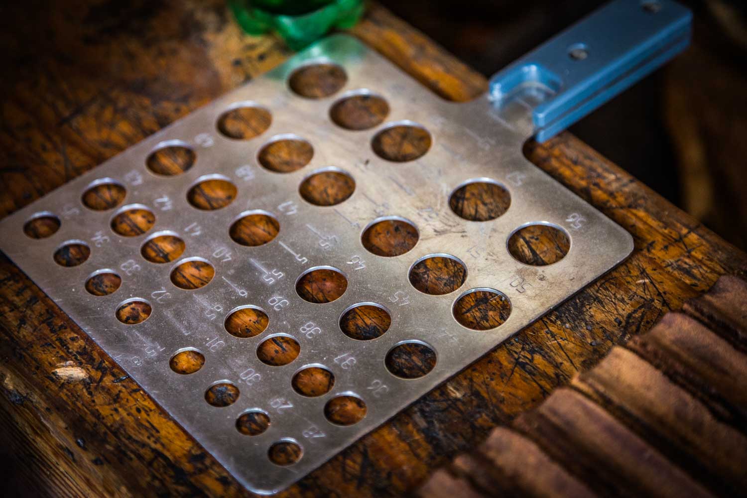 Metal gauge tool with numbered holes on a wooden table, used for measuring wire or drill sizes.