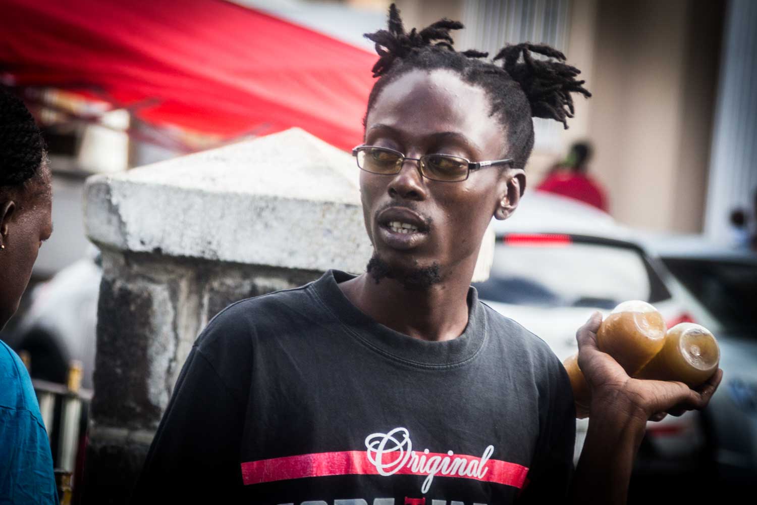 Man in glasses holds bottled drinks at an outdoor market, wearing a t-shirt that says Original.