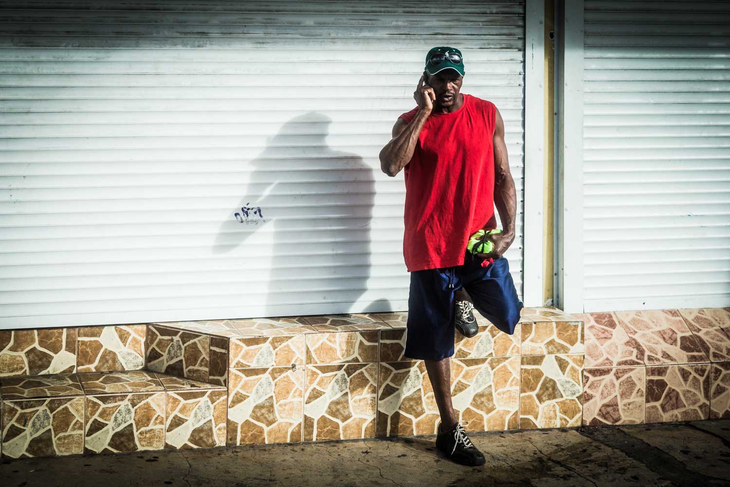Man in red shirt talks on phone, leans against white shutter with mosaic steps.