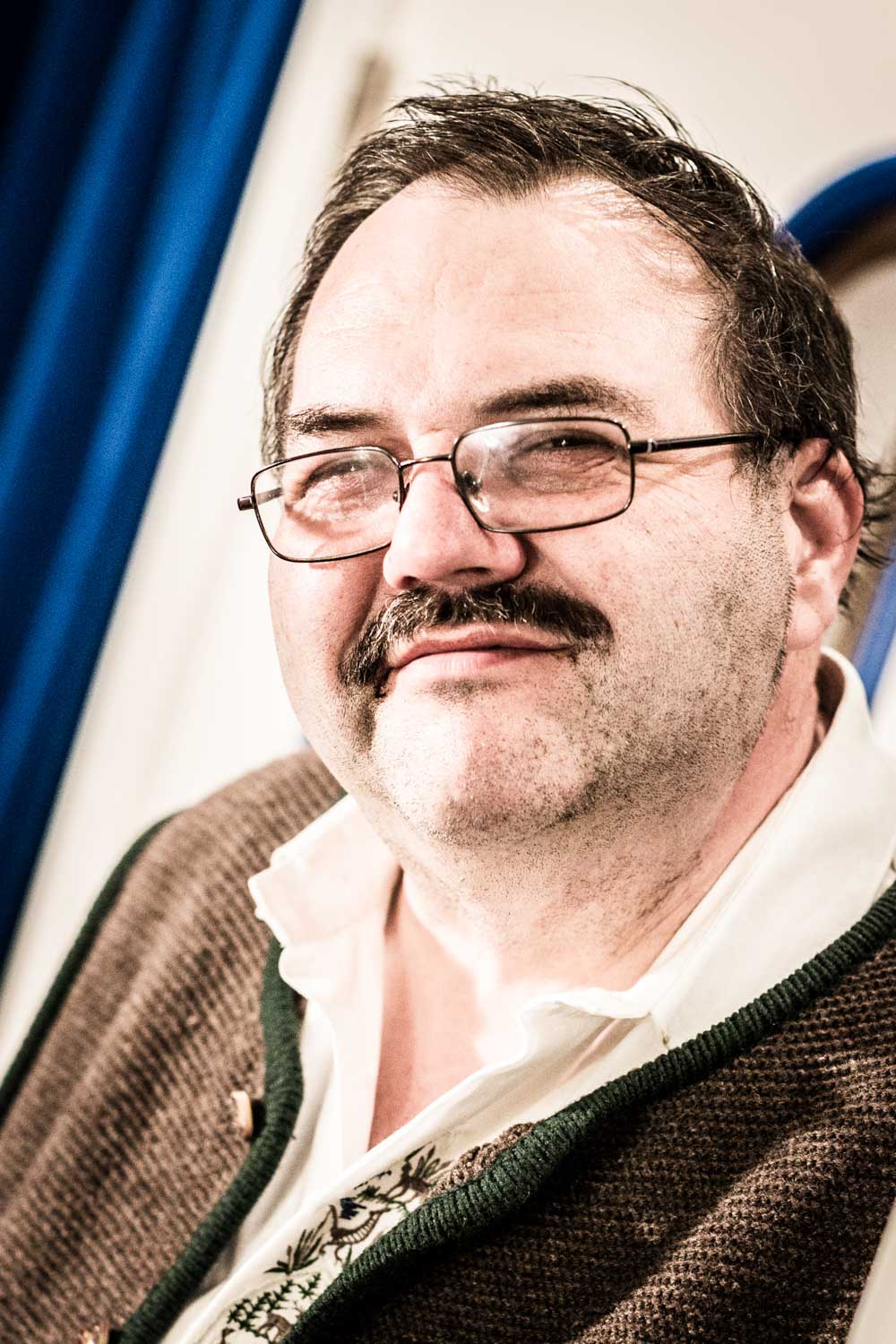 Smiling man with glasses and mustache wearing a traditional style jacket indoors.