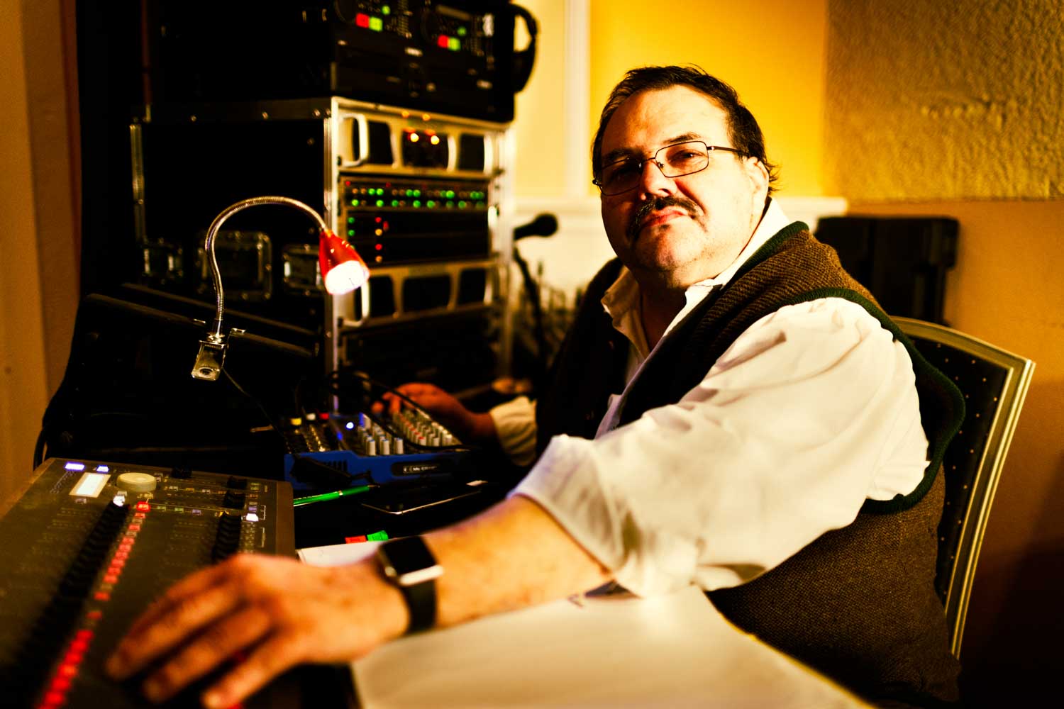 Audio engineer at a sound mixing console with equipment rack in background.