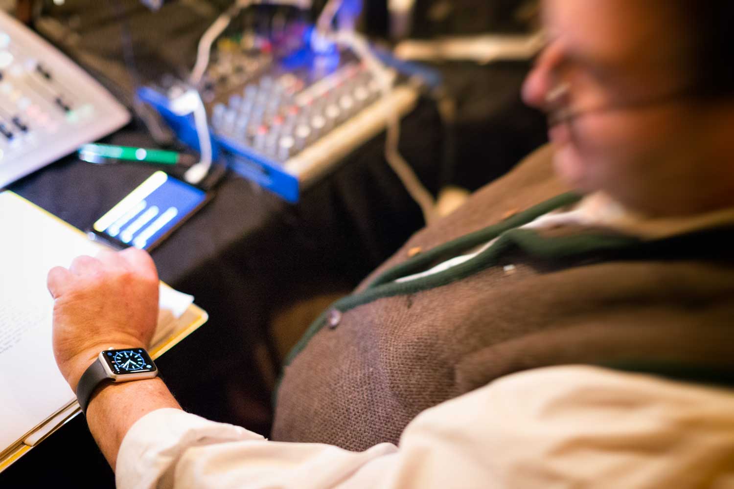 Man checks smartwatch while sitting at mixing console; smartphone and notepad nearby.