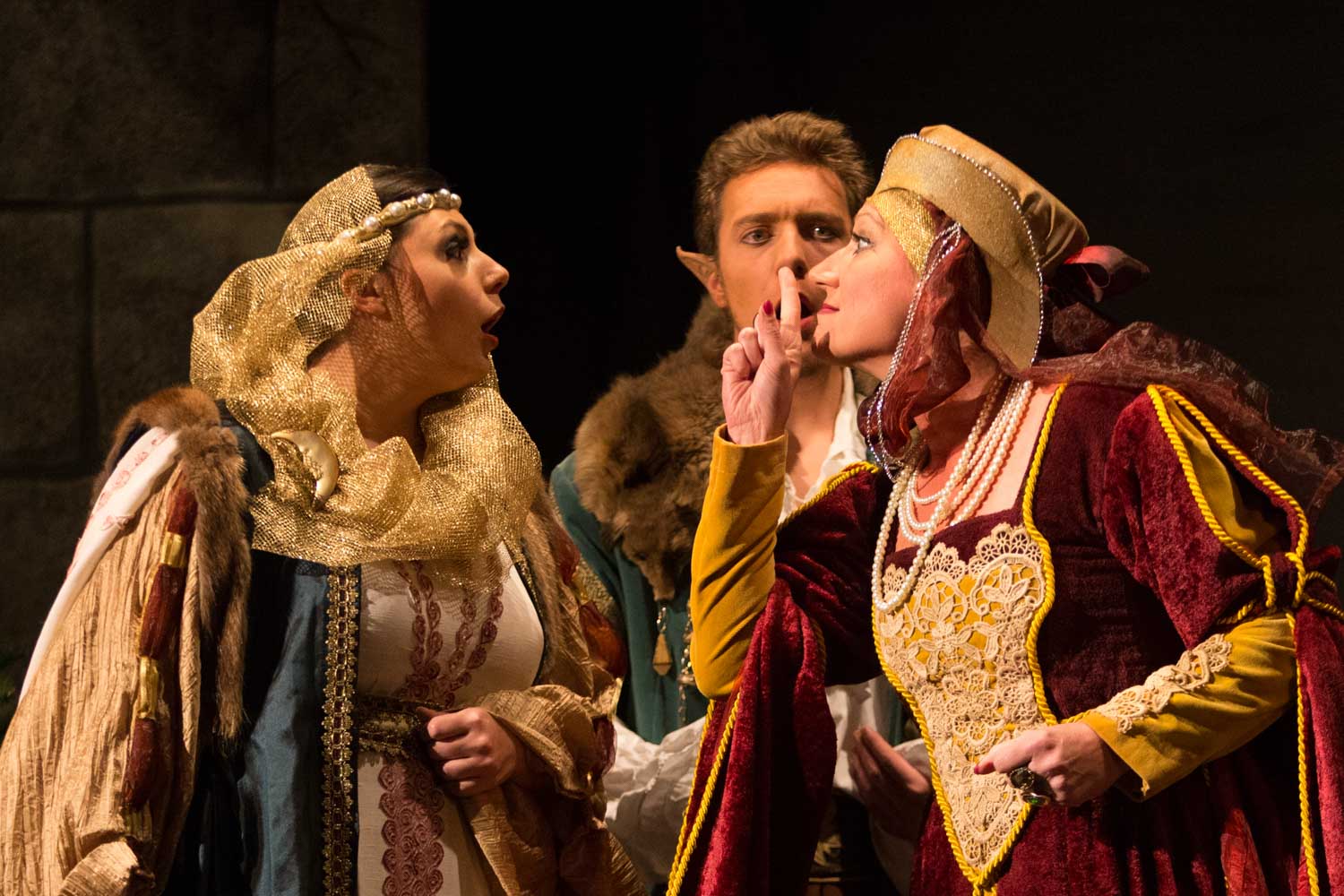 Three actors in medieval costumes perform a dramatic scene on stage, with one gesturing to be quiet.