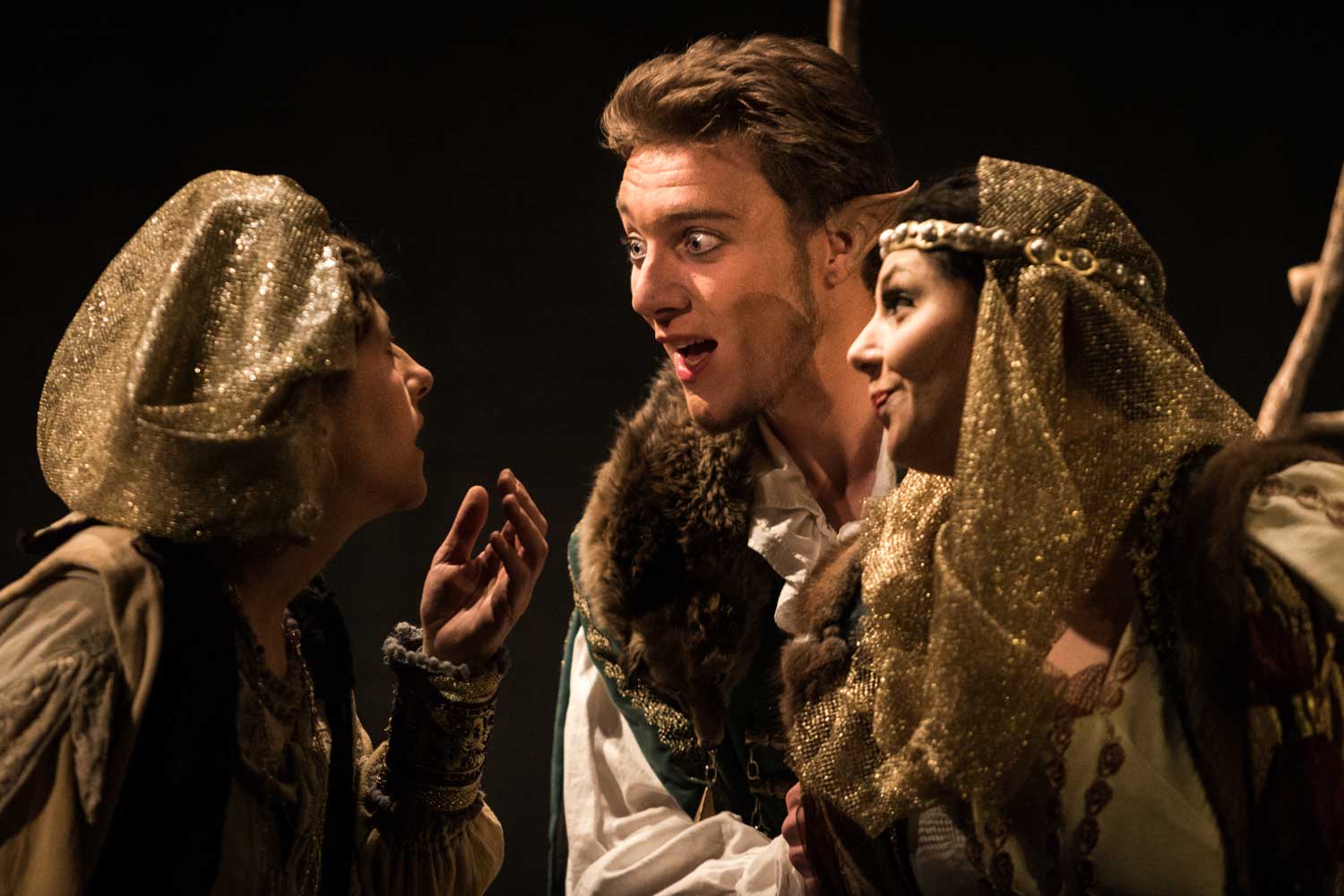 Three actors in medieval costumes perform expressively in a theatrical scene with dim lighting.