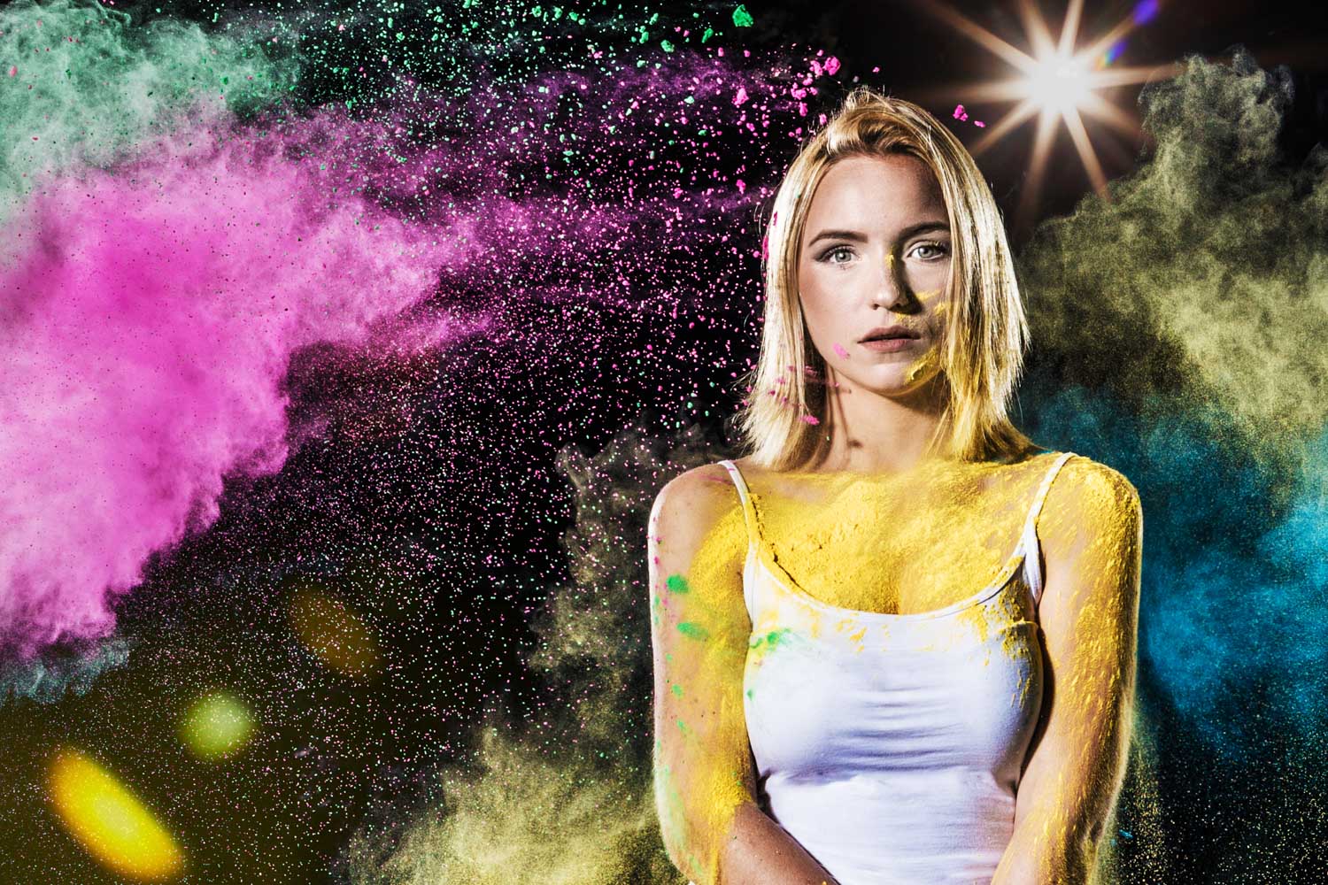 Blonde woman in white tank top with colorful powder on face and body, vibrant pink and green powder in the air.